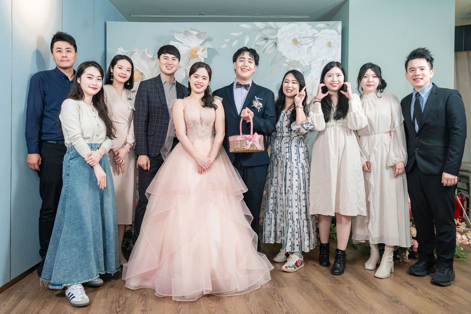 Wedding party group poses together in front of a floral backdrop wearing formal attire in pastel colors.