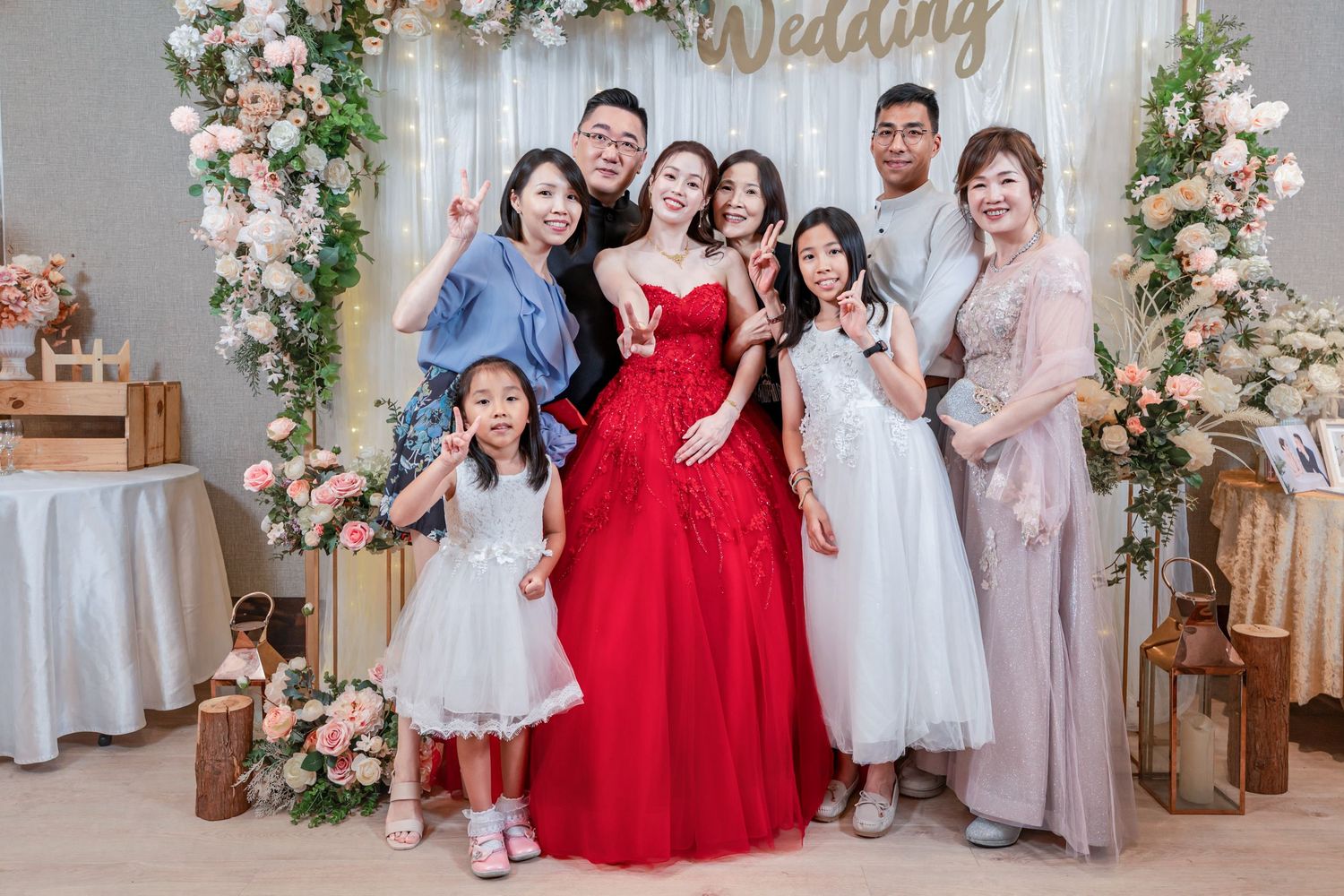 Wedding party in red and neutral dresses poses together with floral decorations and rustic wooden elements.