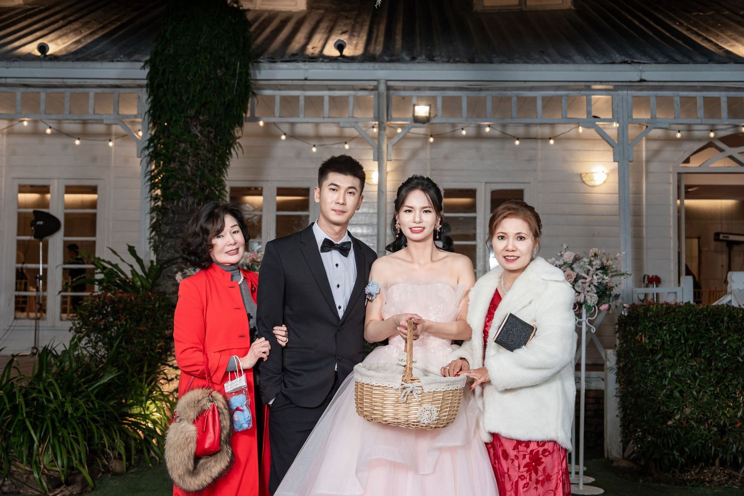 Wedding party poses together in front of an elegant venue with glass windows and greenery at dusk.