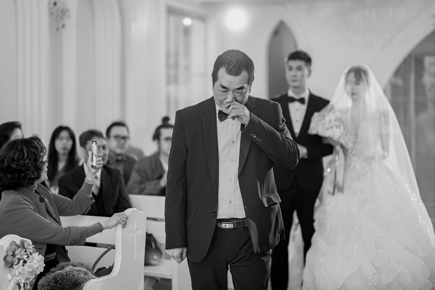 Black and white photo of a wedding ceremony in progress with attendees seated in rows and a person speaking in a suit.