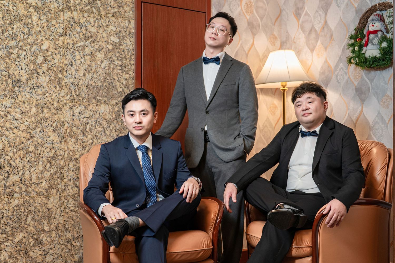 Three well-dressed men in formal suits and bow ties pose together on brown leather furniture in an elegant setting.