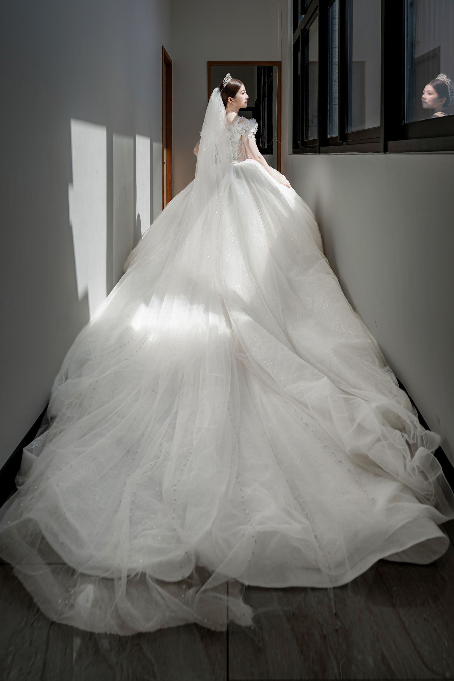 Bride in a voluminous white tulle ball gown with a long train and veil stands in a sunlit hallway, viewed from behind.