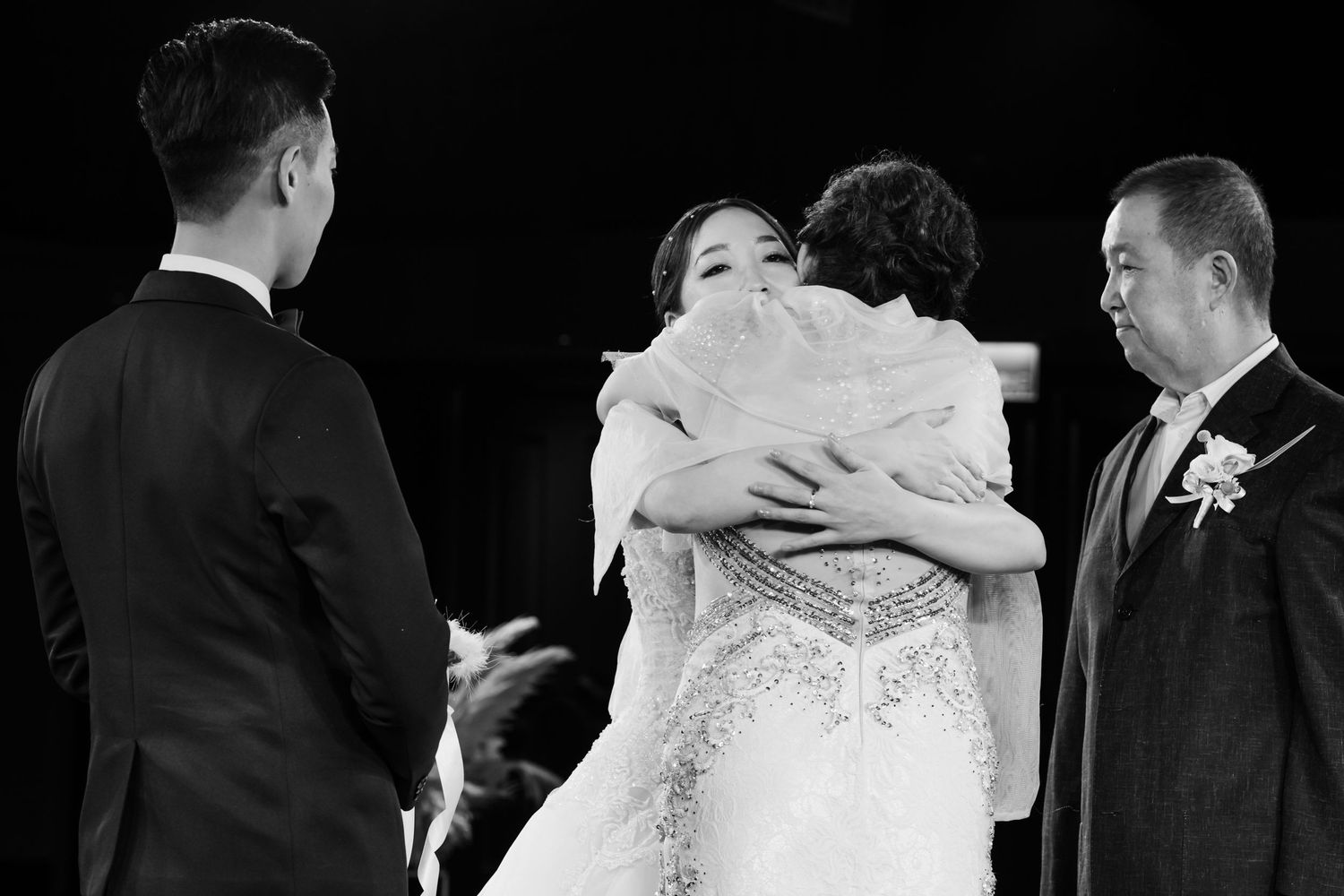 A black and white wedding ceremony moment showing a bride in a sparkly gown embracing someone while others look on.
