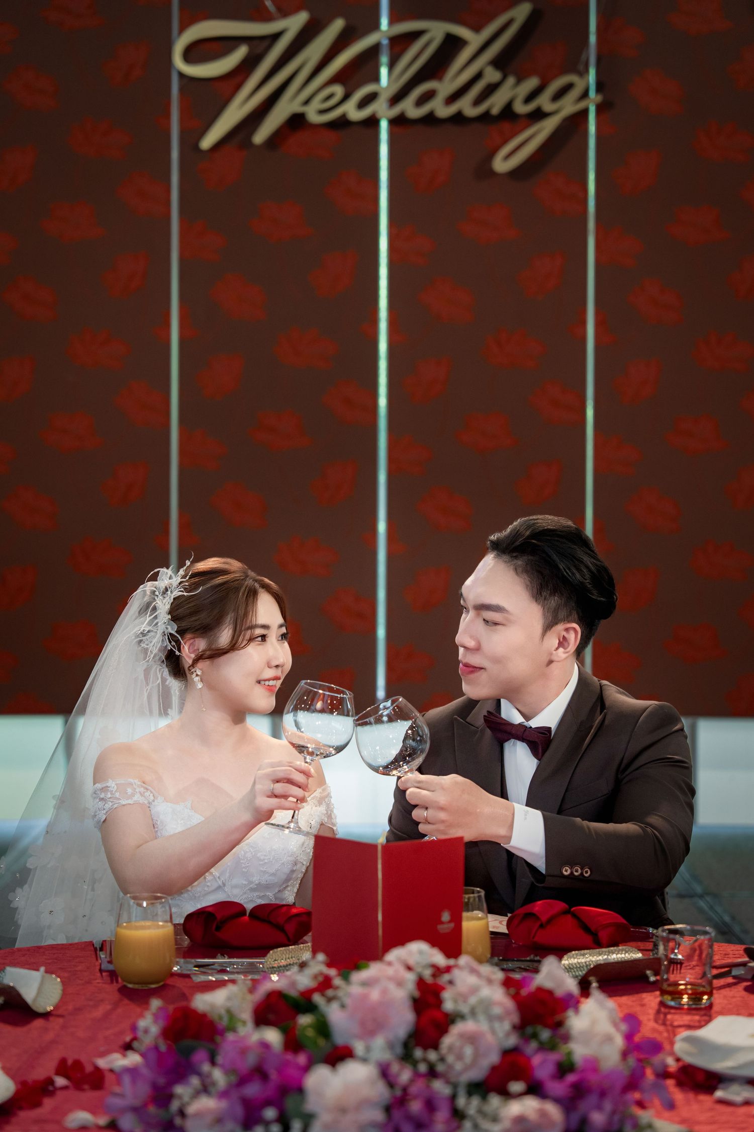 A couple in wedding attire shares drinks and a romantic moment at their reception table decorated with colorful flowers.