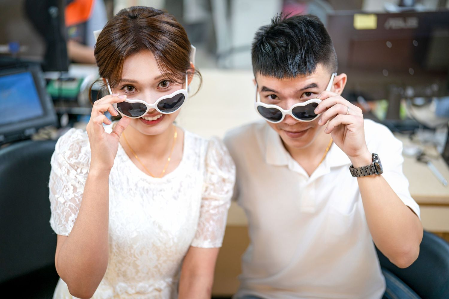 Two people in white outfits and sunglasses sit together at a table outdoors on a sunny day.