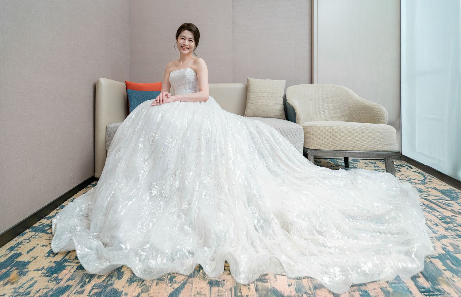 A person wearing a flowing white lace wedding dress lies gracefully on a decorative carpet floor in a bright room.