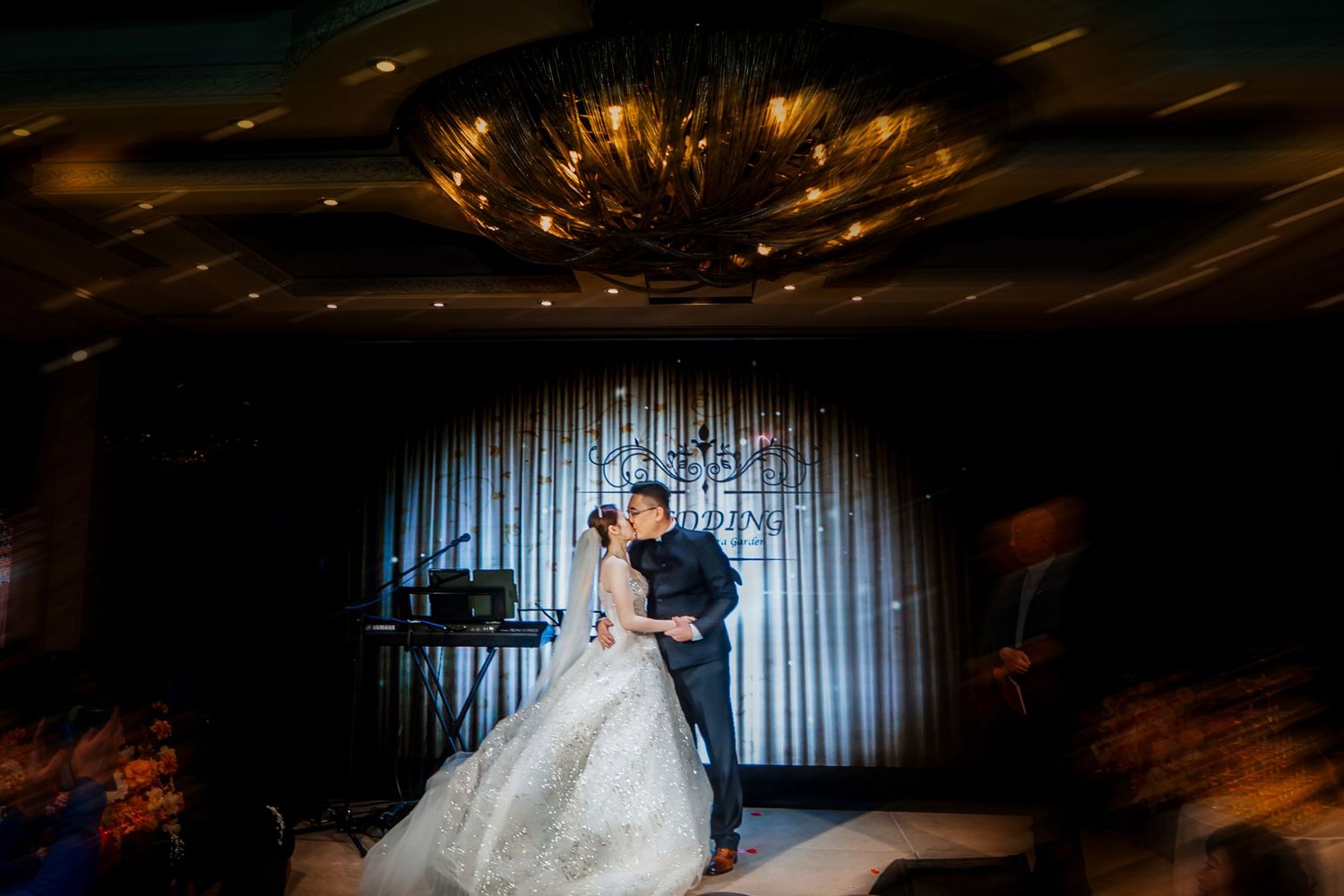 A couple in wedding attire share a romantic dance in a dimly lit venue with dramatic lighting.