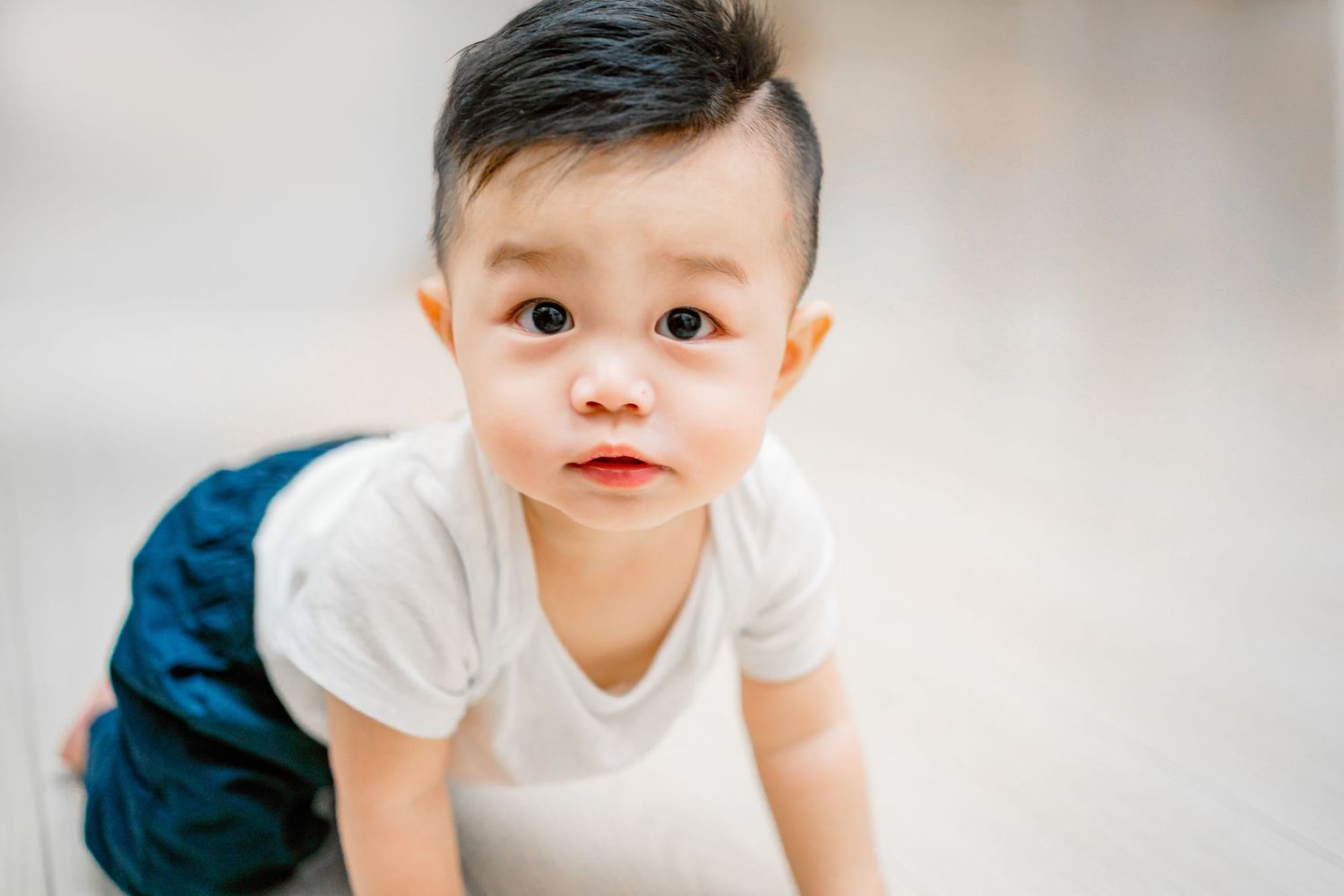 A baby in a white shirt and blue pants sits and plays while looking at the camera with curiosity.