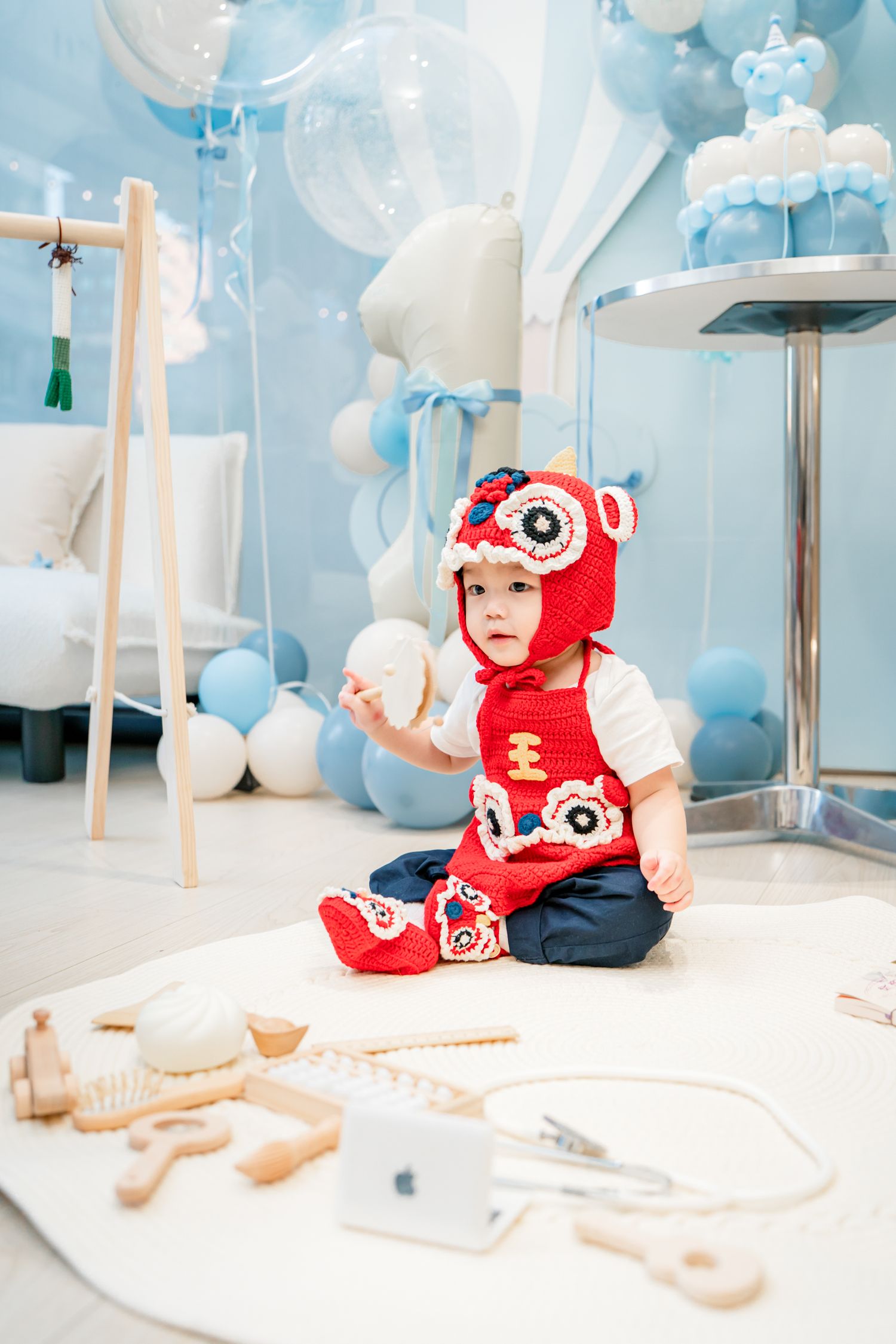 Baby in red outfit plays on floor with wooden toys under dreamy blue and white cloud-themed party decorations.
