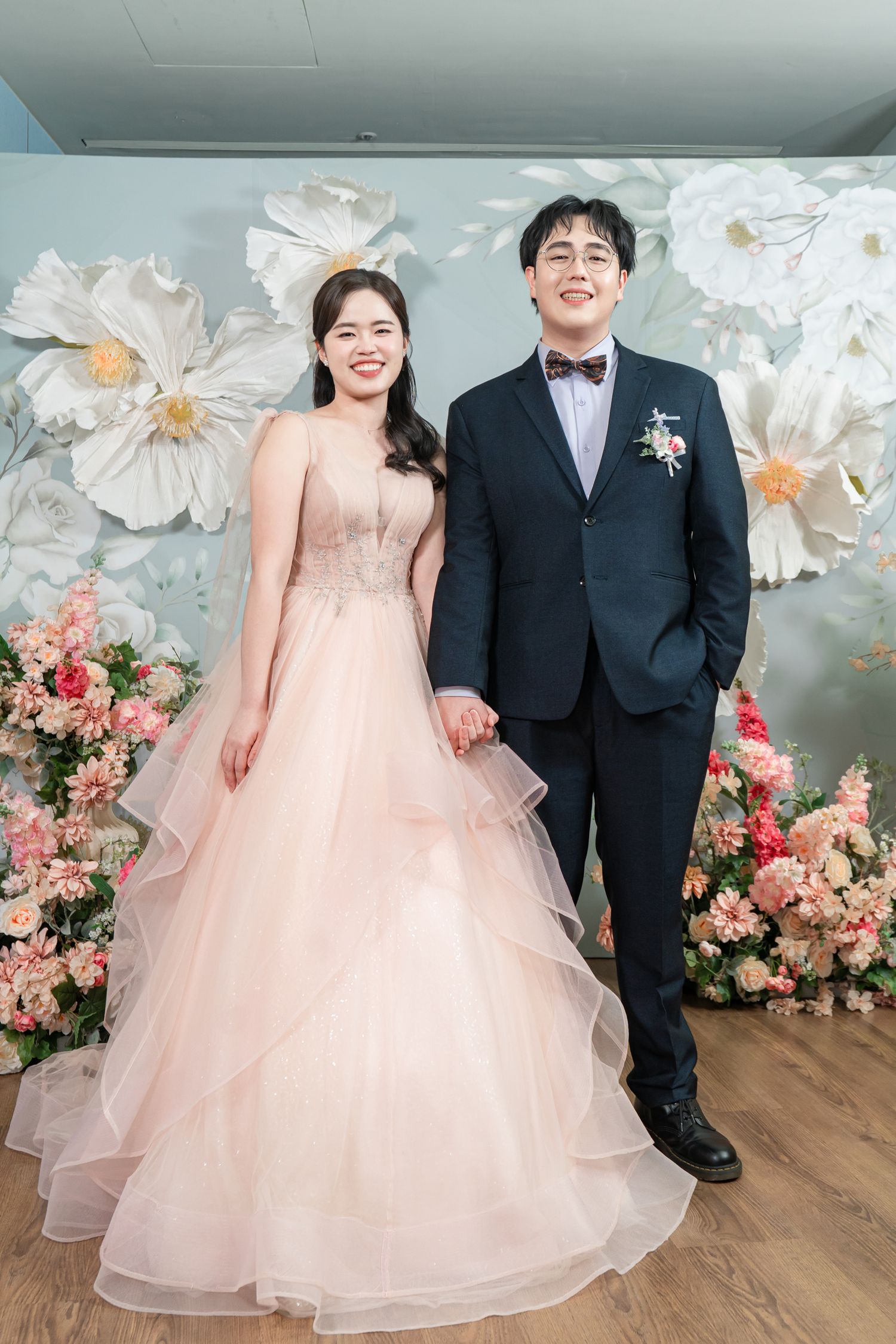 A couple in formal wedding attire stands together against a floral backdrop in soft pink and white tones.