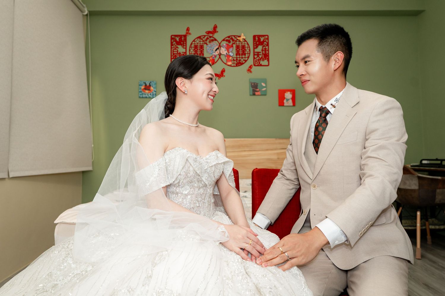 Bride in white gown and groom in beige suit holding hands and smiling at each other in a decorated room.