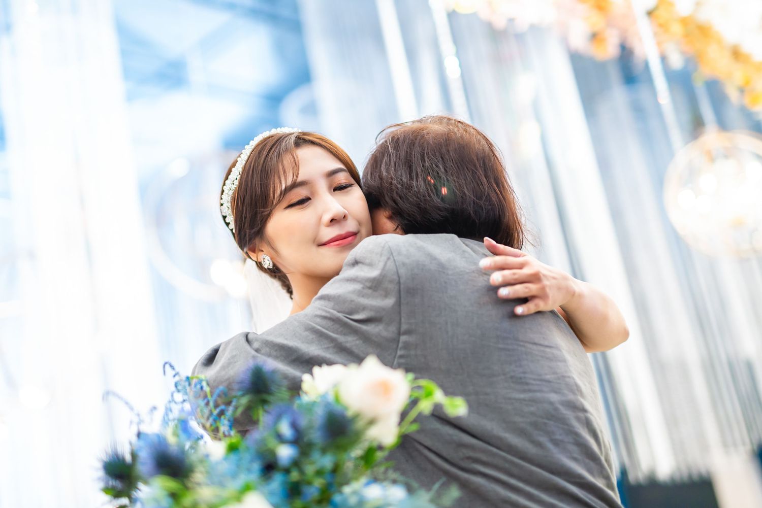 Two people share a warm embrace with flowers in the foreground and a blurred cityscape background.