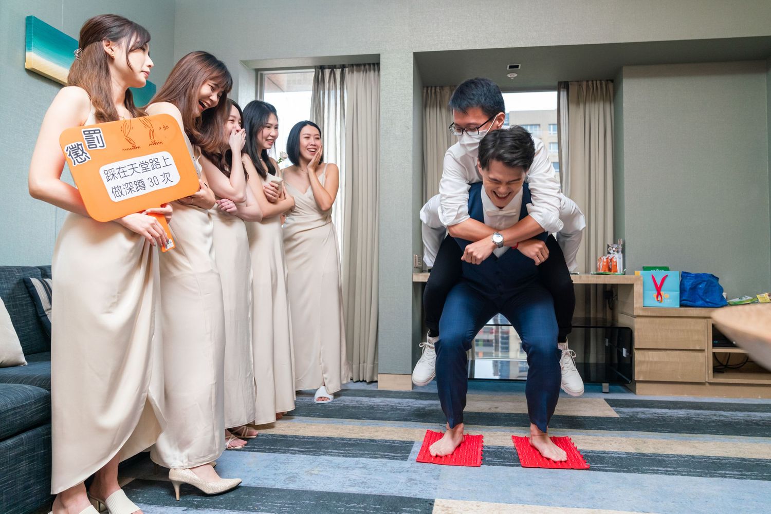 Bridesmaids in beige dresses gather around to watch someone trying on bright red shoes in a living room.
