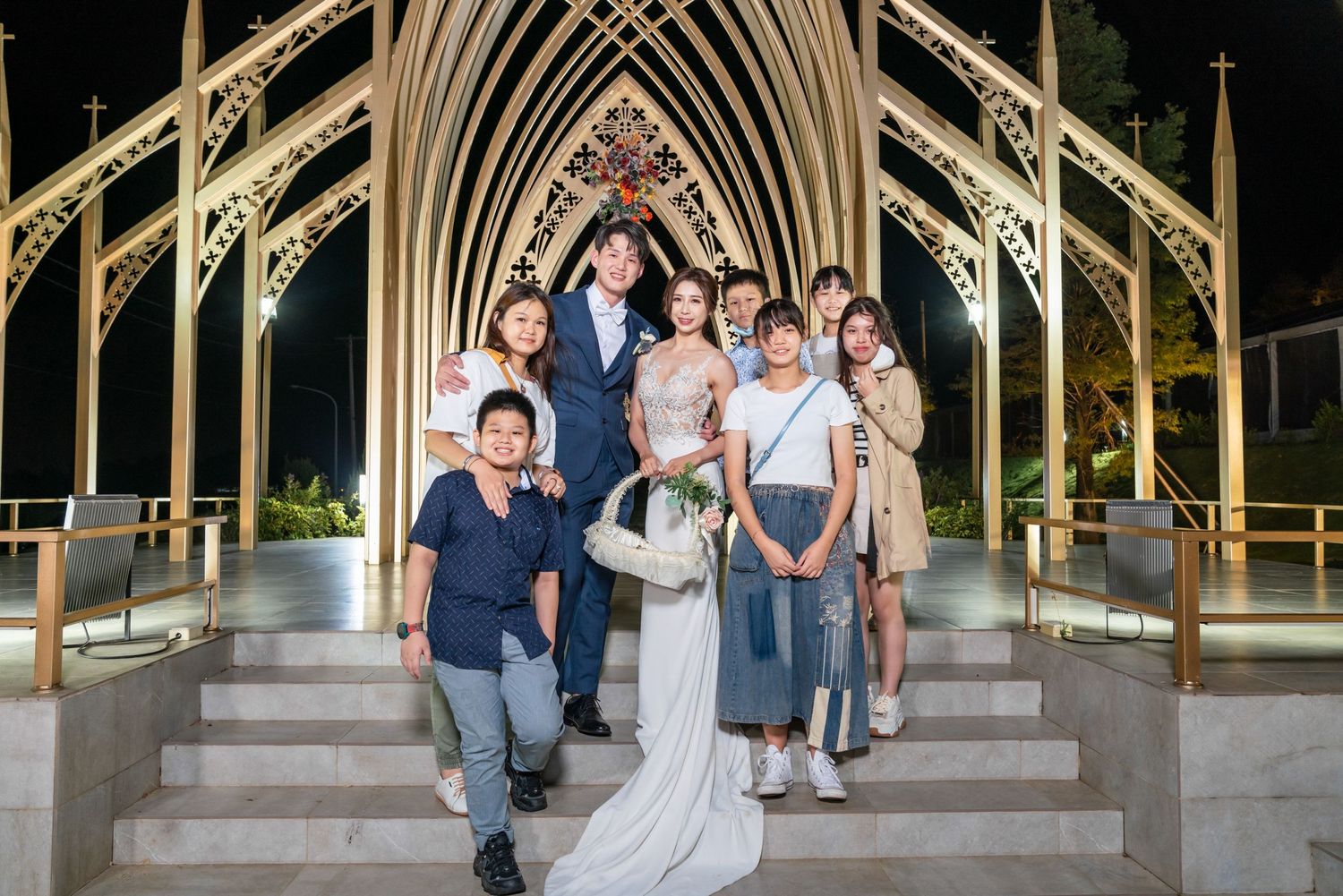 Group poses on elegant stone steps under a Gothic arch with ornate architectural details at night.