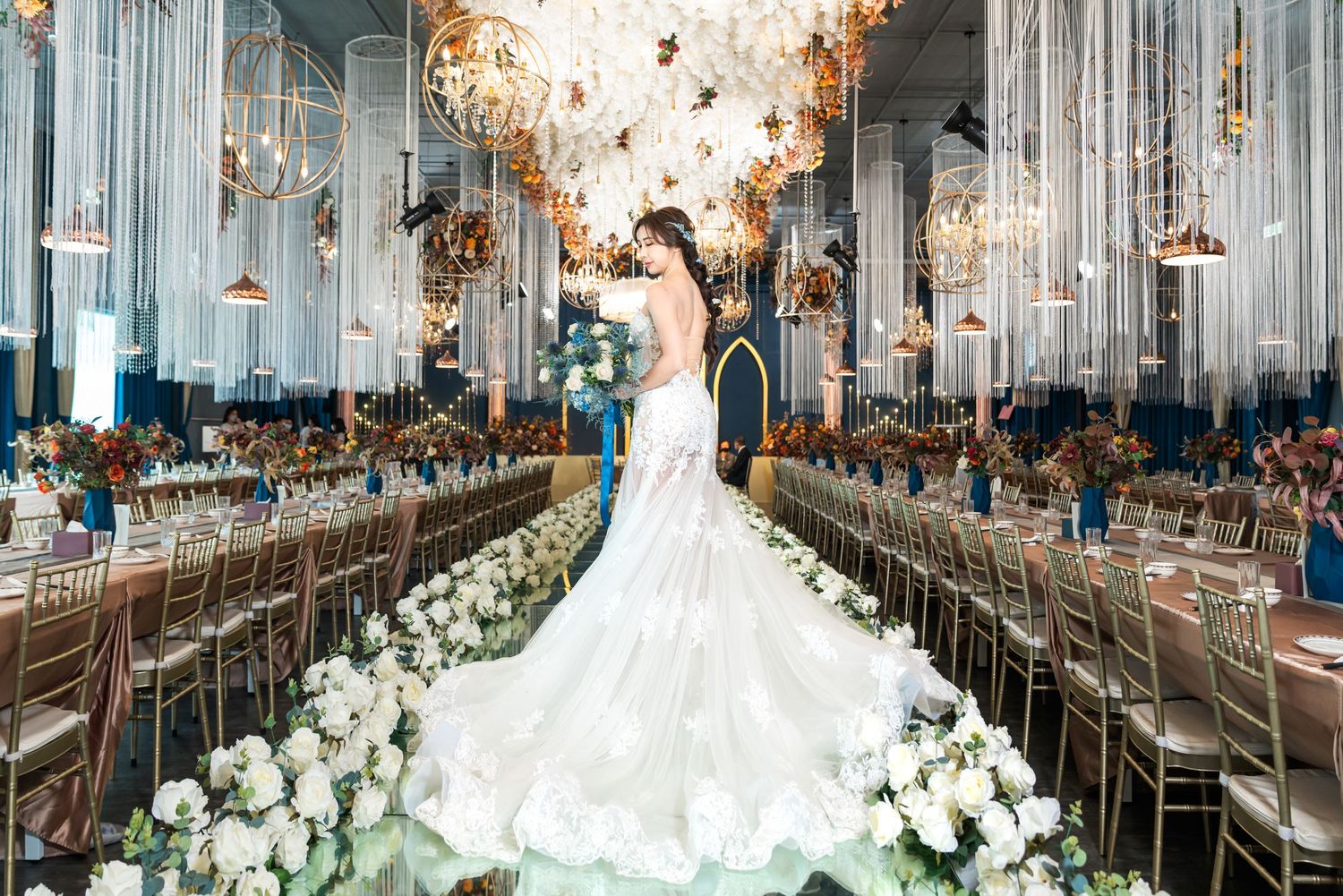 Elegant wedding reception setup in grand hall with crystal chandeliers, white floral arrangements and mirror-lined tables.