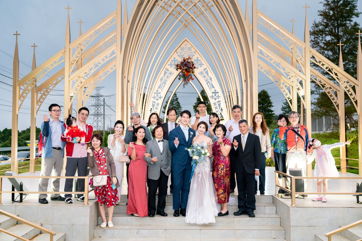Wedding party poses under an elegant white outdoor chapel with arched beams and clear sky background.