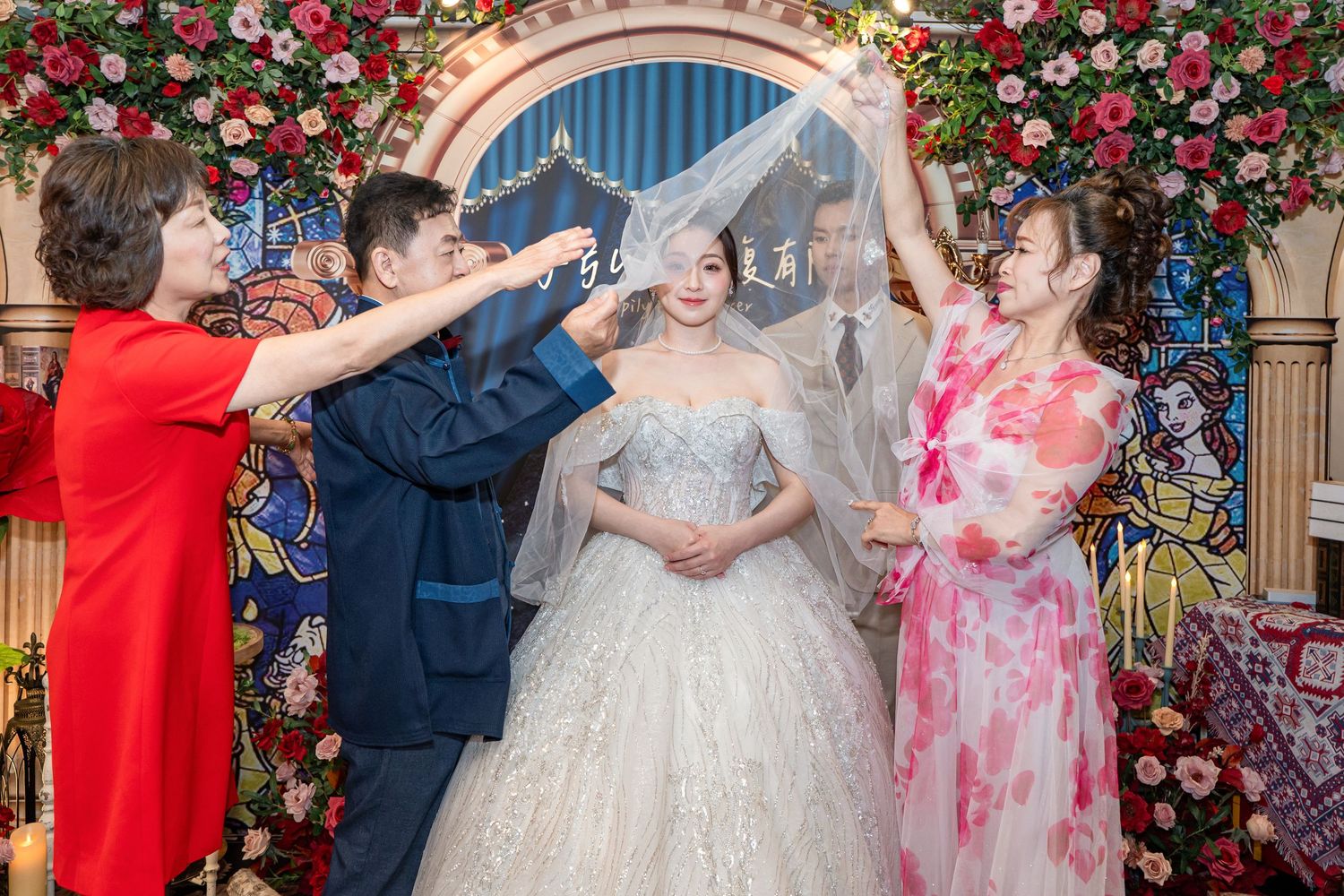 A groom lifts a bridal veil over his bride in a floral wedding ceremony setting with family members assisting.