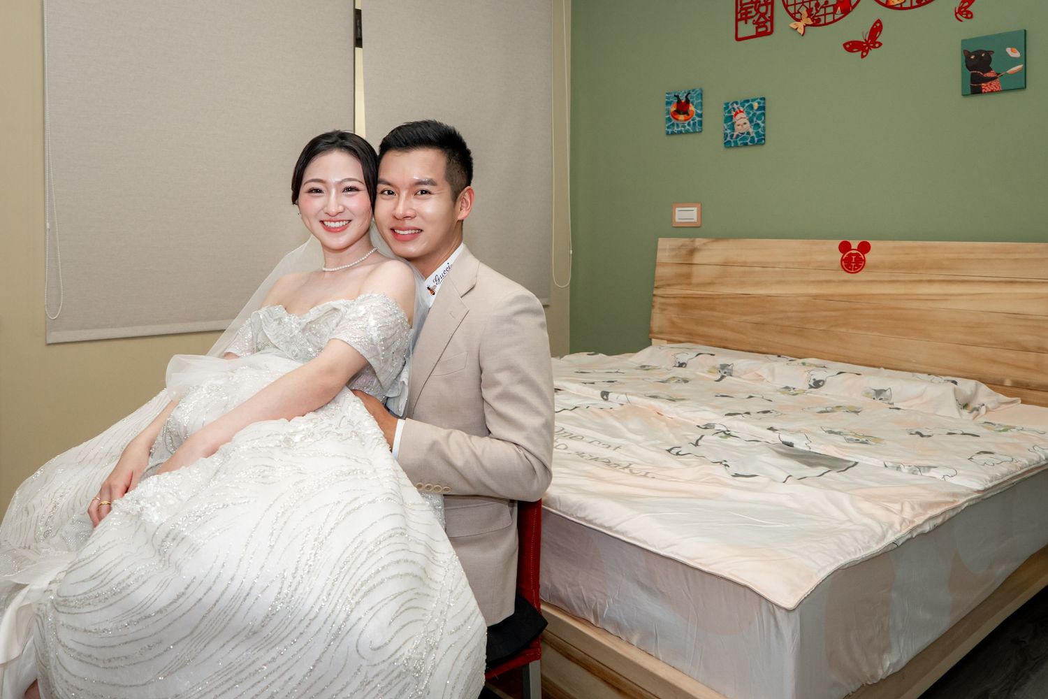 A couple in wedding attire poses elegantly beside a bed, the bride in a white gown and groom in a beige suit.
