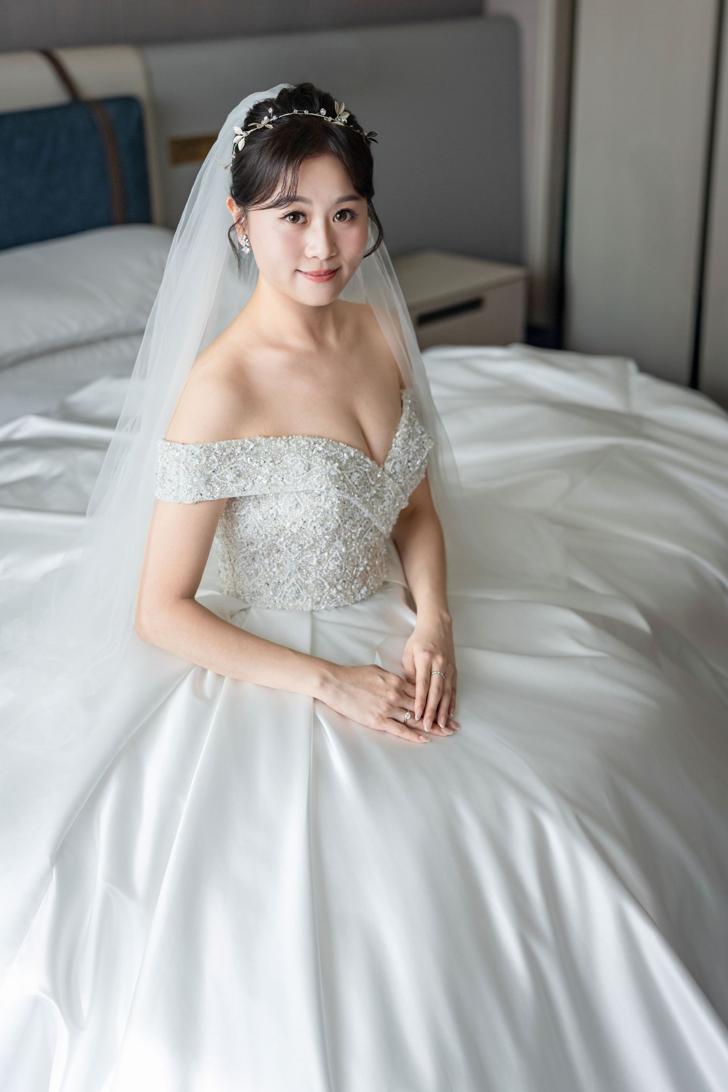 A bride in an elegant off-shoulder white ball gown with beaded bodice and flowing veil sits on white bedding.