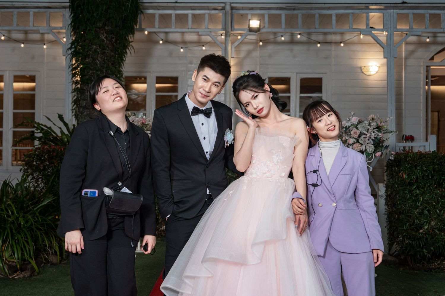 A wedding party poses together outdoors in formal attire including a pink gown and suits in front of a lit building.