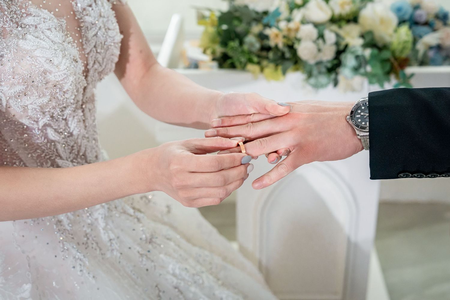 Close-up of hands exchanging wedding rings during a ceremony with white lace dress and floral decorations.