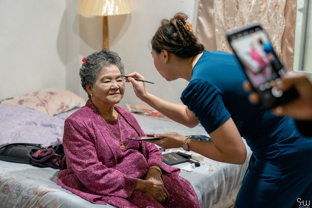 A makeup artist applies cosmetics to someone wearing a pink robe during wedding preparations.