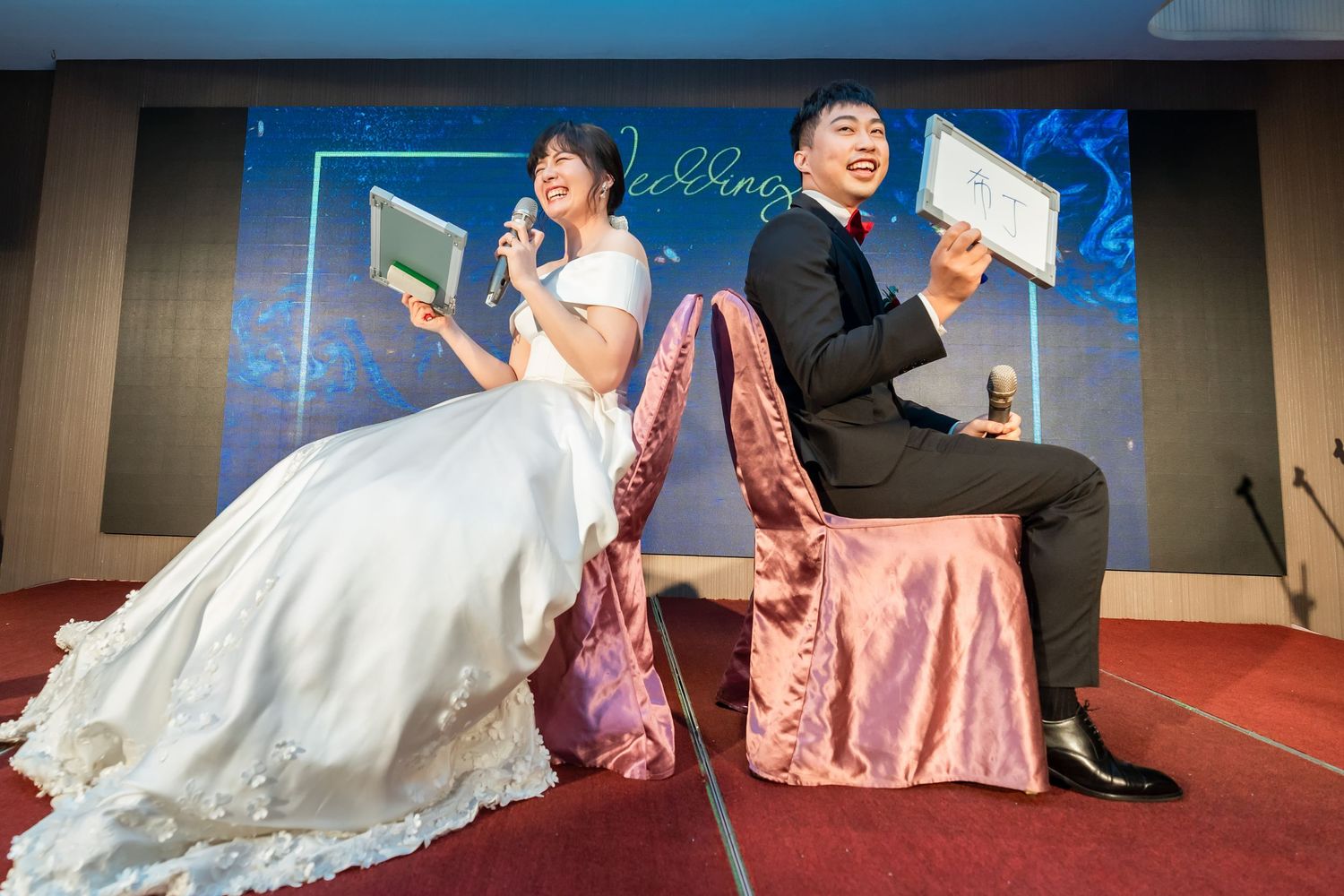 A newlywed couple sits on pink velvet chairs at their wedding reception holding papers.
