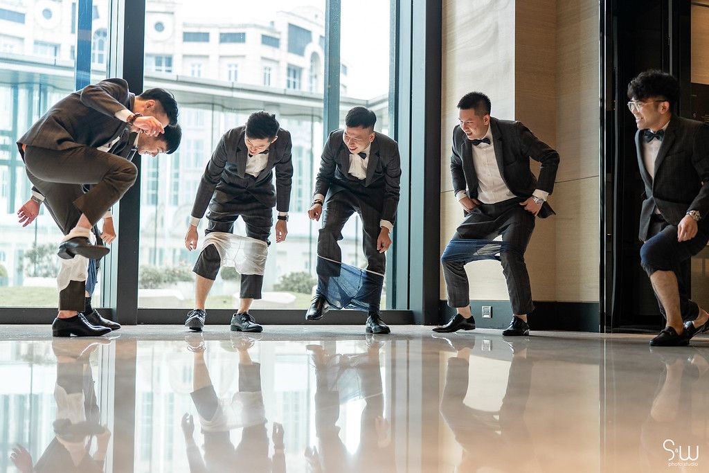 Groomsmen in formal wear line up while bending down in a modern building hallway.