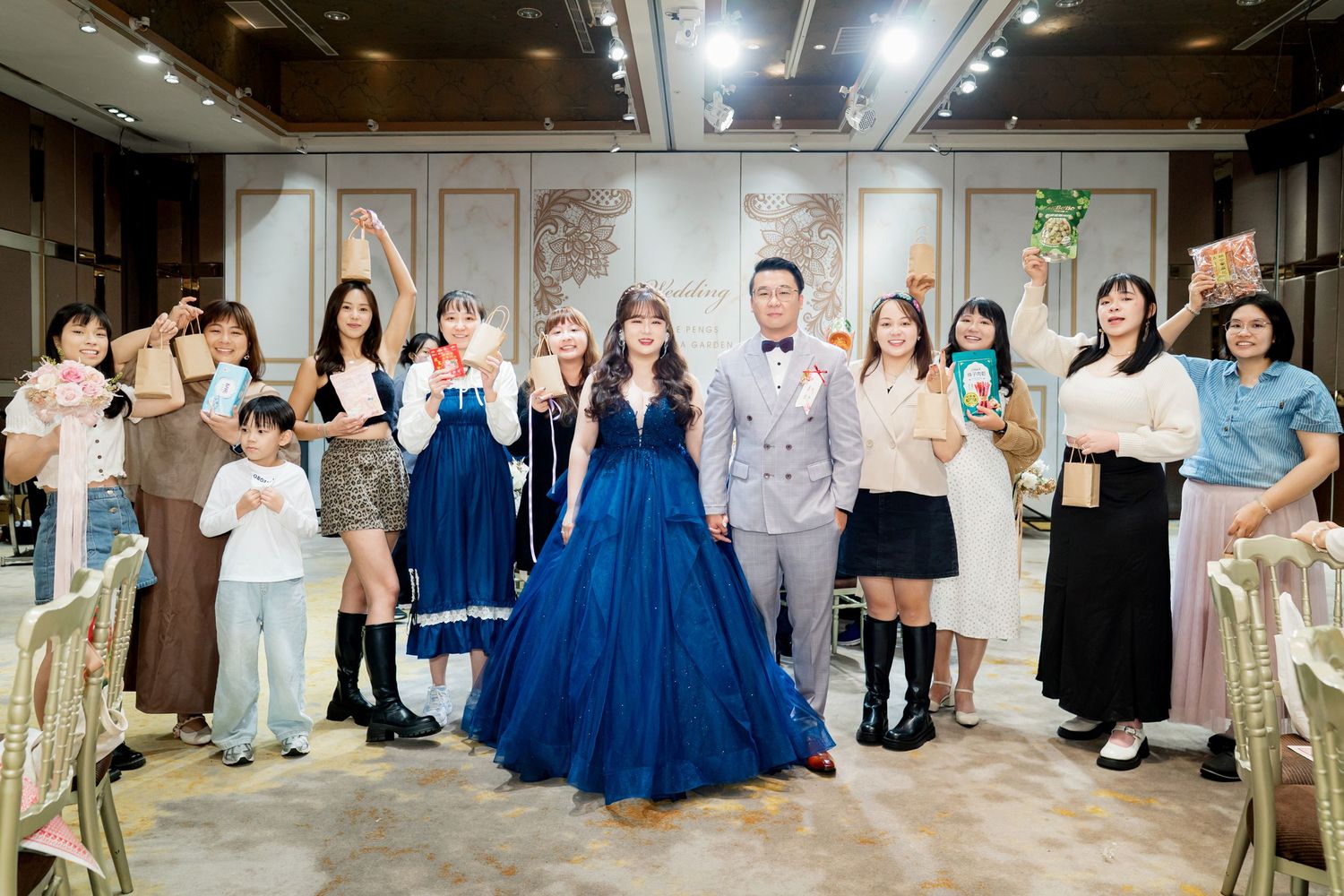 Wedding party guests in formal attire celebrate together in a spacious reception hall with festive poses and expressions.