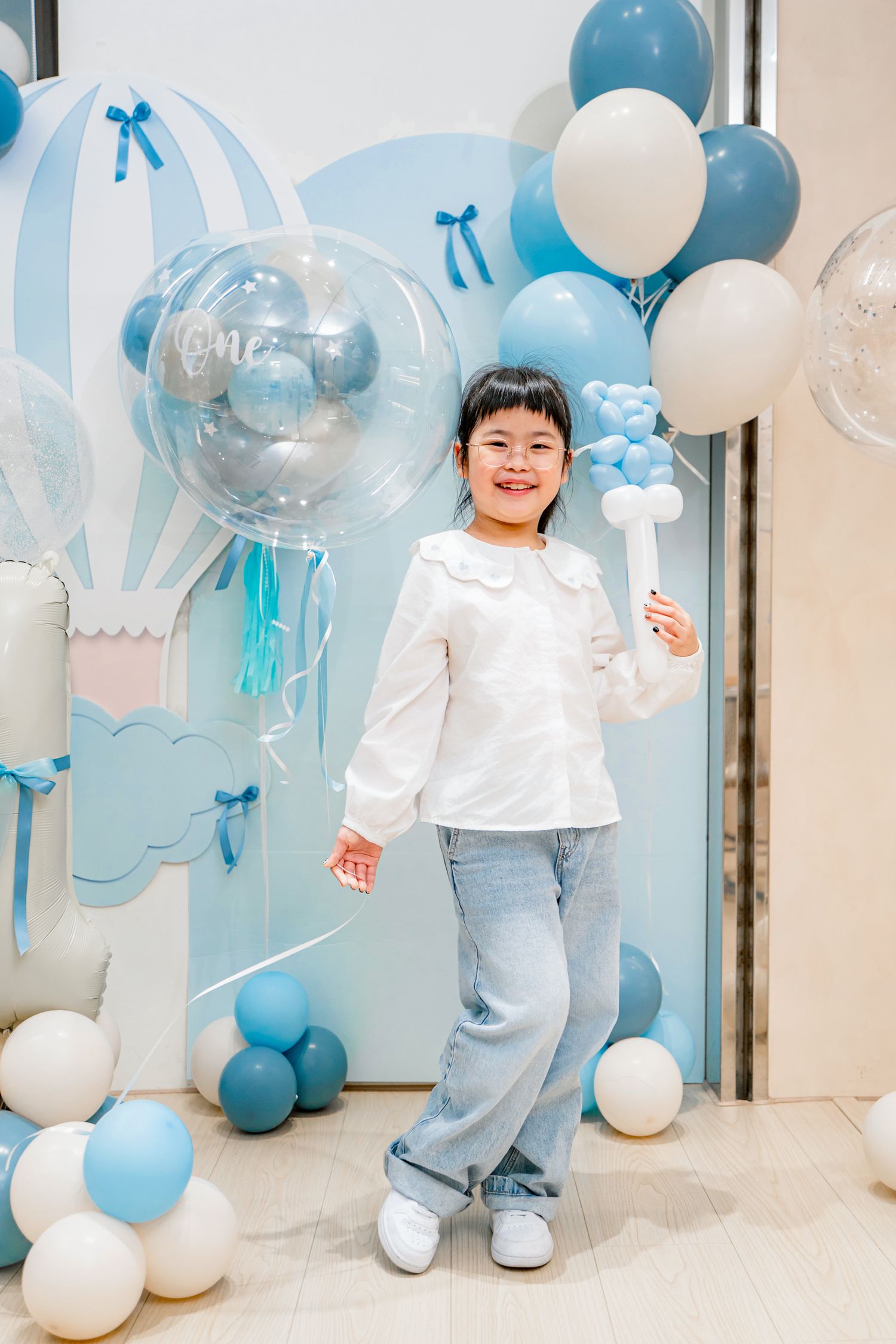 Festive party setup with blue and white balloons, bows, and decorative elements against a light colored wall.