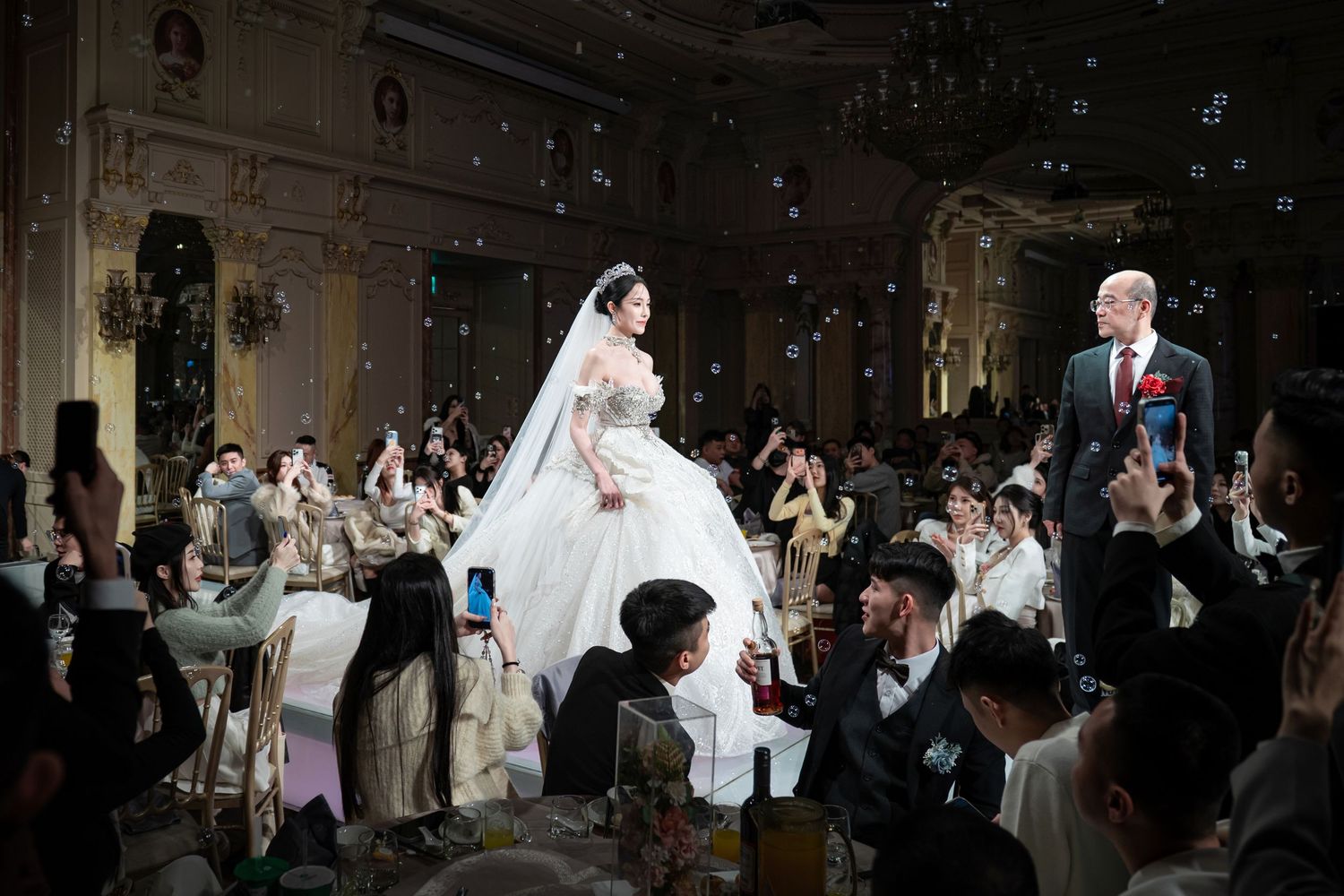 Wedding guests celebrate during a joyous reception in an elegant ballroom with ornate decor and floating bubbles.