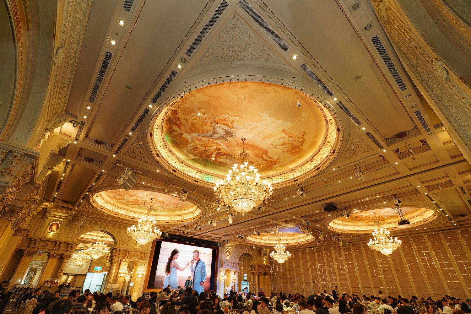 Elegant ballroom with ornate ceiling murals, crystal chandeliers and large projection screen during an event.