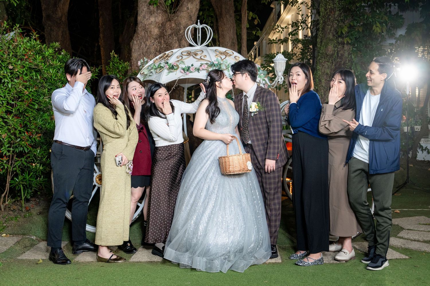 A group of wedding guests pose together outdoors at night under decorative lighting.