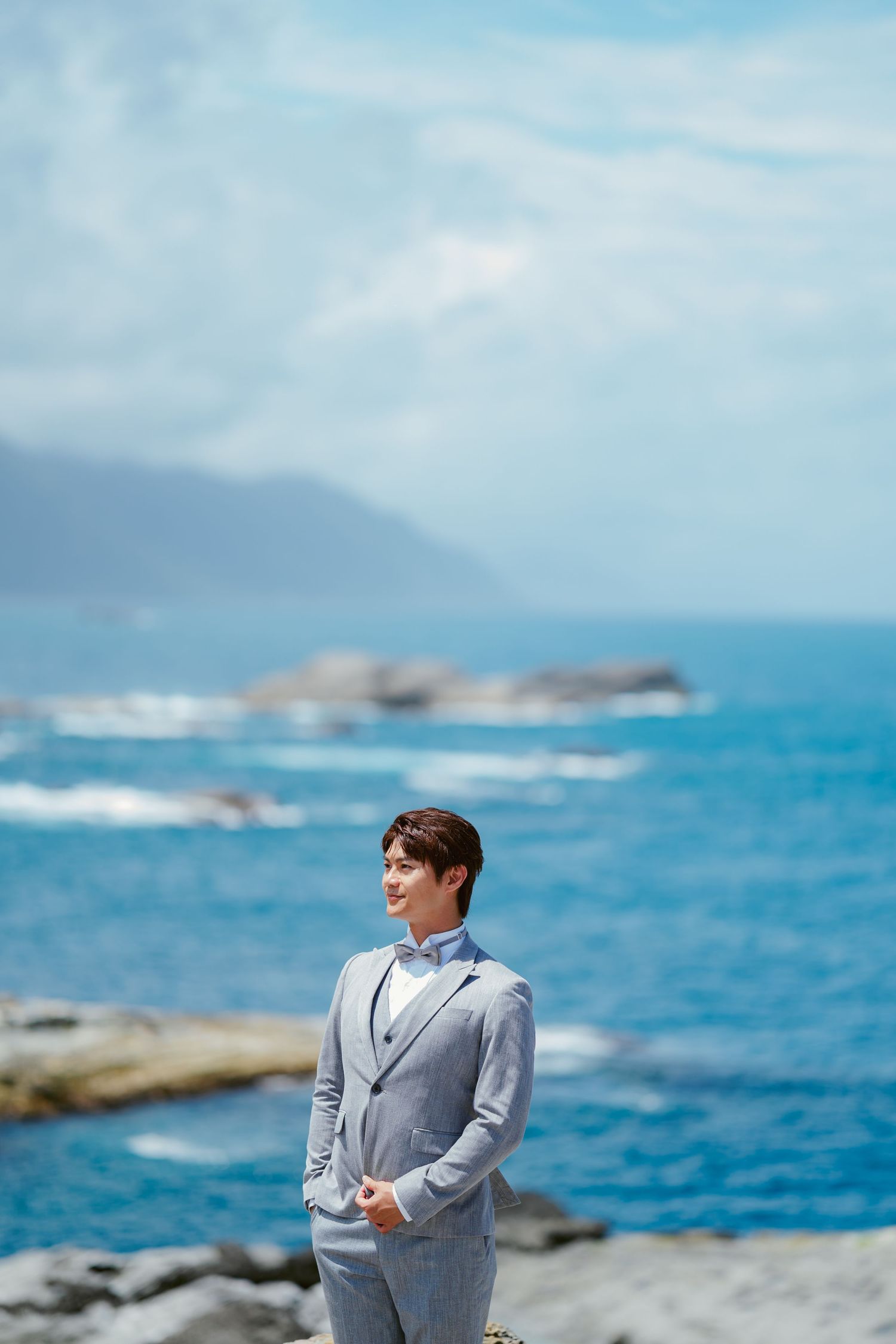 Person in light gray suit stands on rocky coastline with bright blue ocean and cloudy sky in the background.