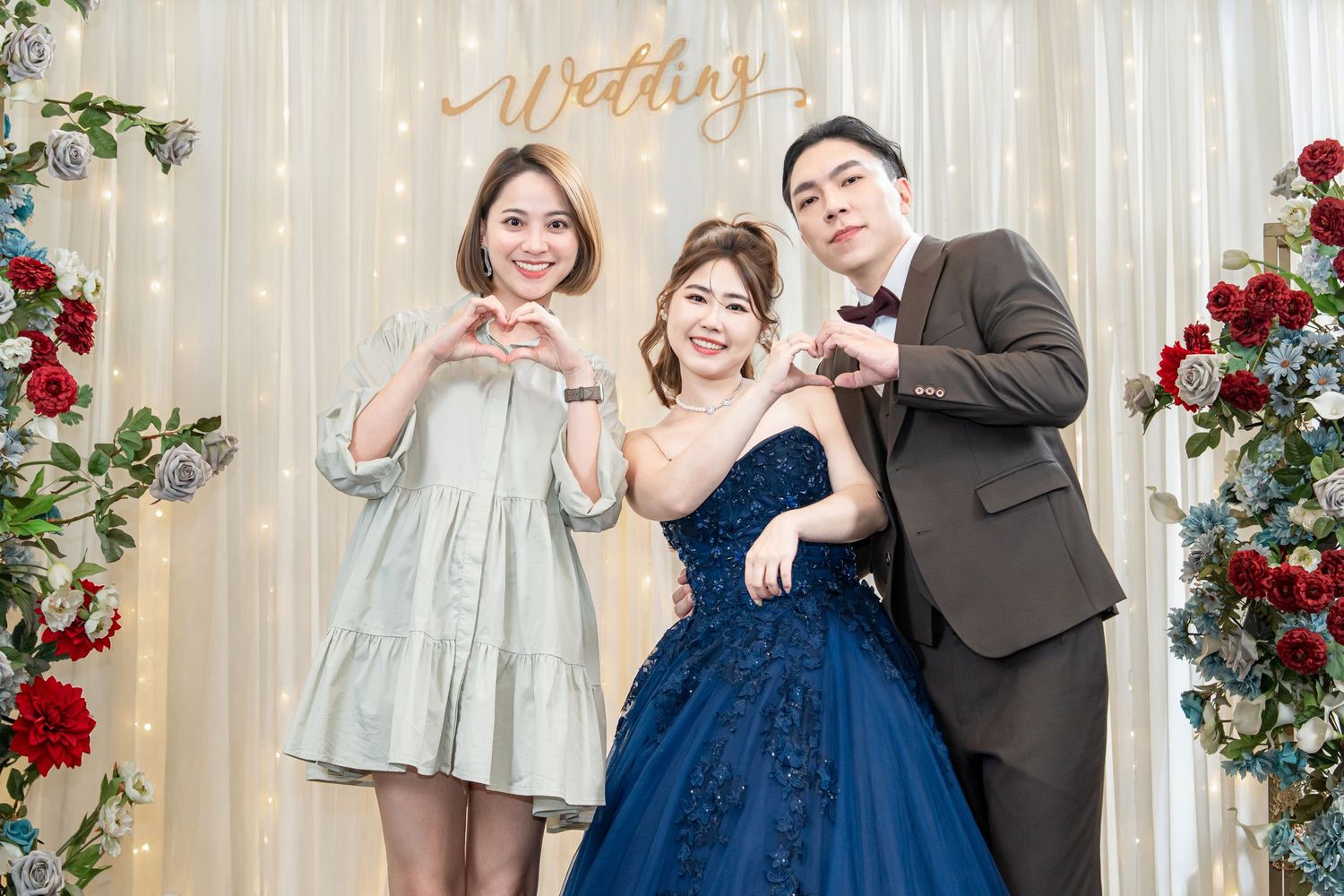 Three people pose together at a wedding photo backdrop with white curtains and red rose decorations.