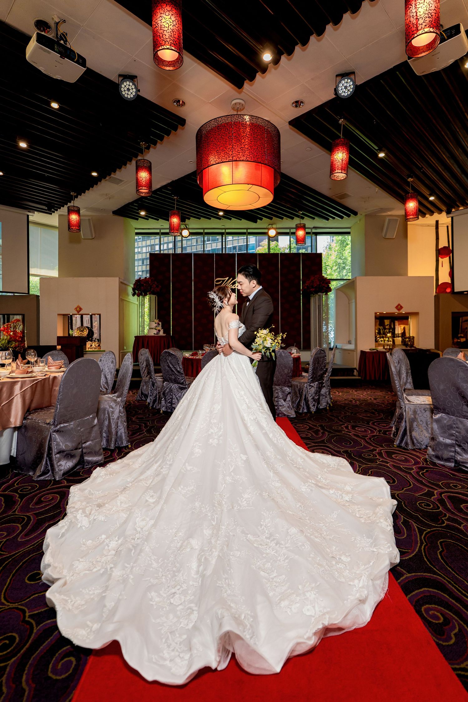 A stunning white wedding gown with a dramatic cathedral train spreads across a red carpeted venue with Asian lanterns.