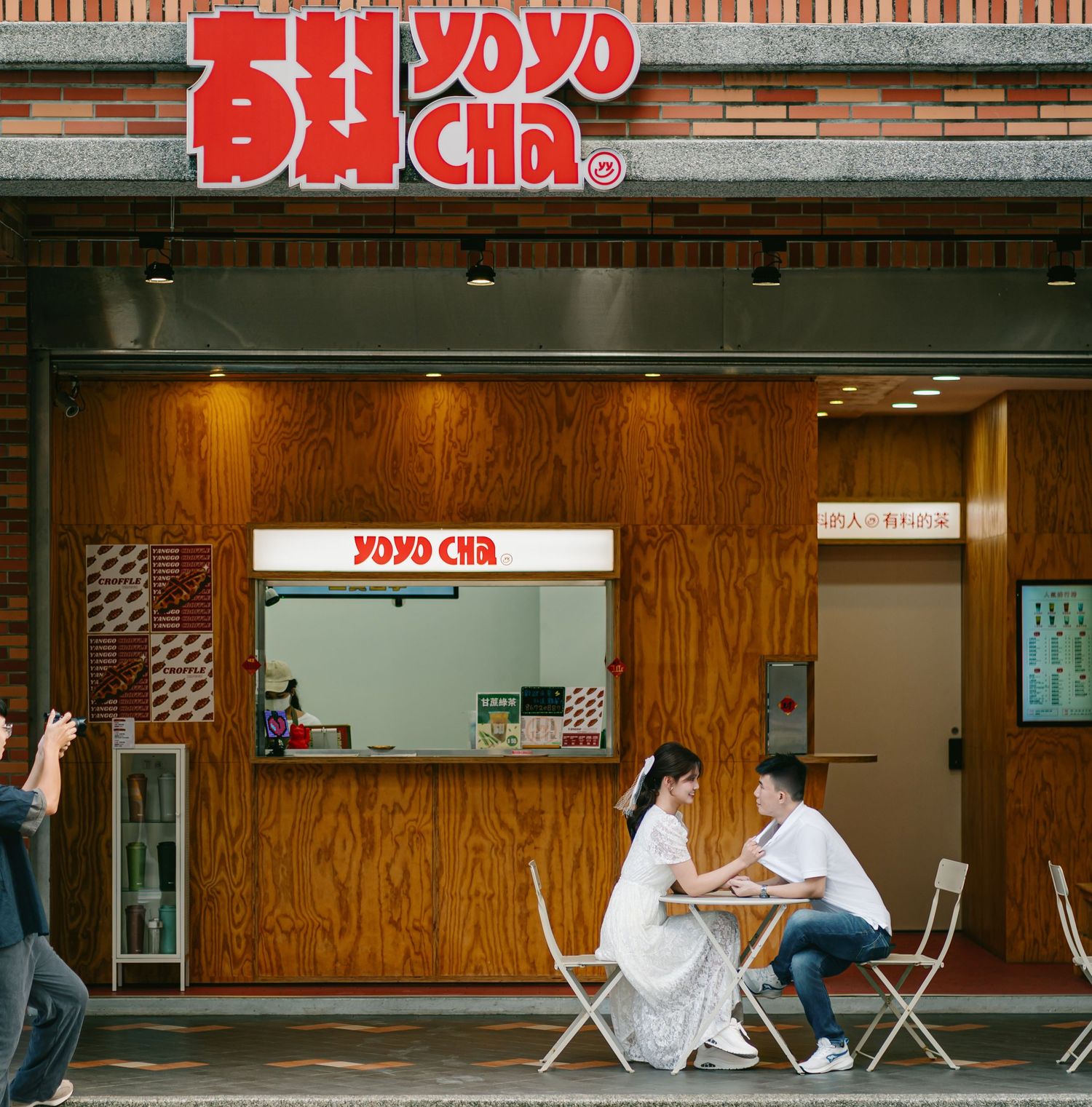 A couple sits at a small table outside a wood-paneled Yoyo Cha cafe storefront with retro signage.