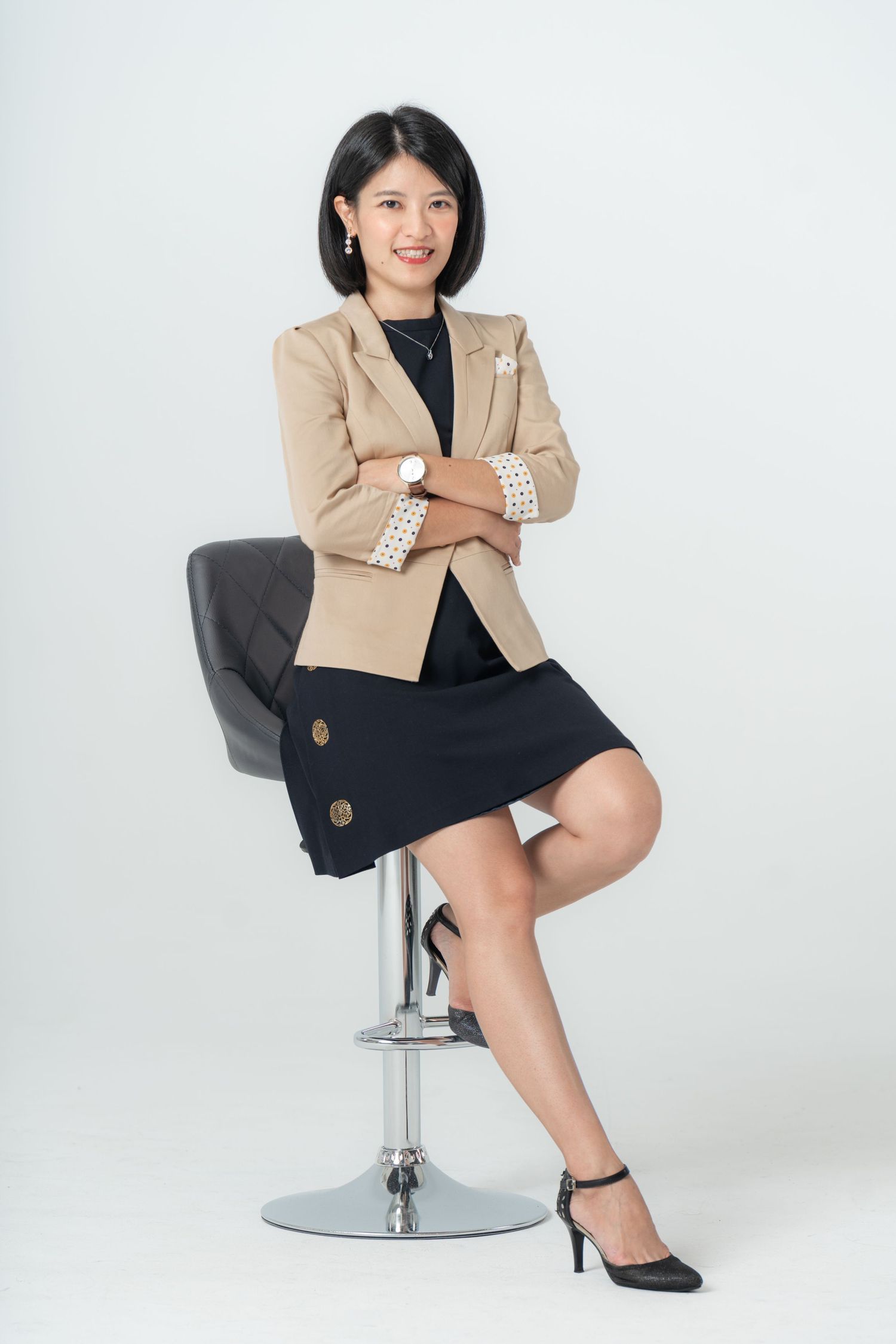 Professional woman in beige blazer and black skirt poses confidently on modern gray swivel chair in studio setting.