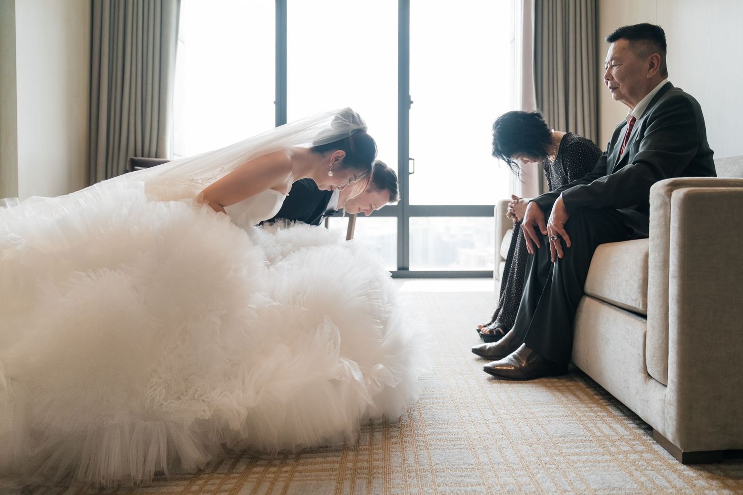 A bride in a flowing white dress lounges on a beige sofa in a bright hotel room.