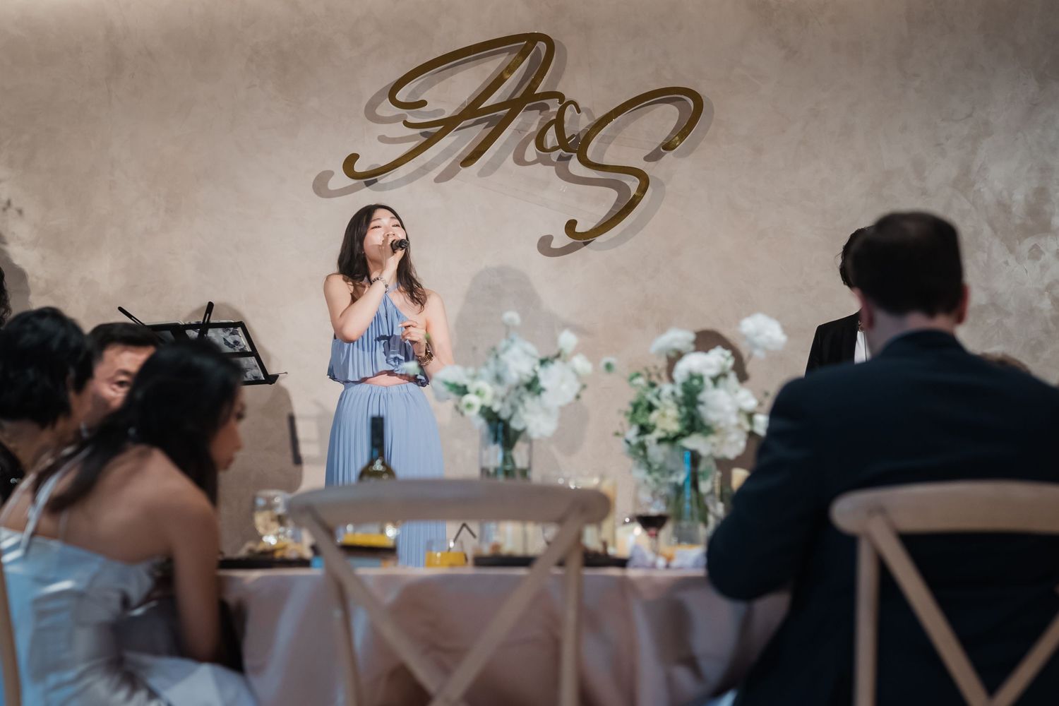 Wedding reception guest delivers emotional speech at decorated table with floral centerpieces and gold monogram backdrop.
