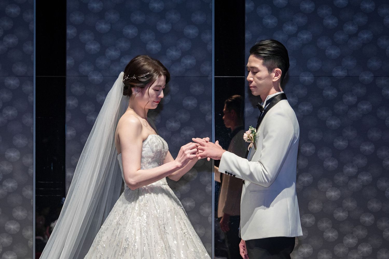 A bride and groom exchange rings during a wedding ceremony against a blue textured backdrop.
