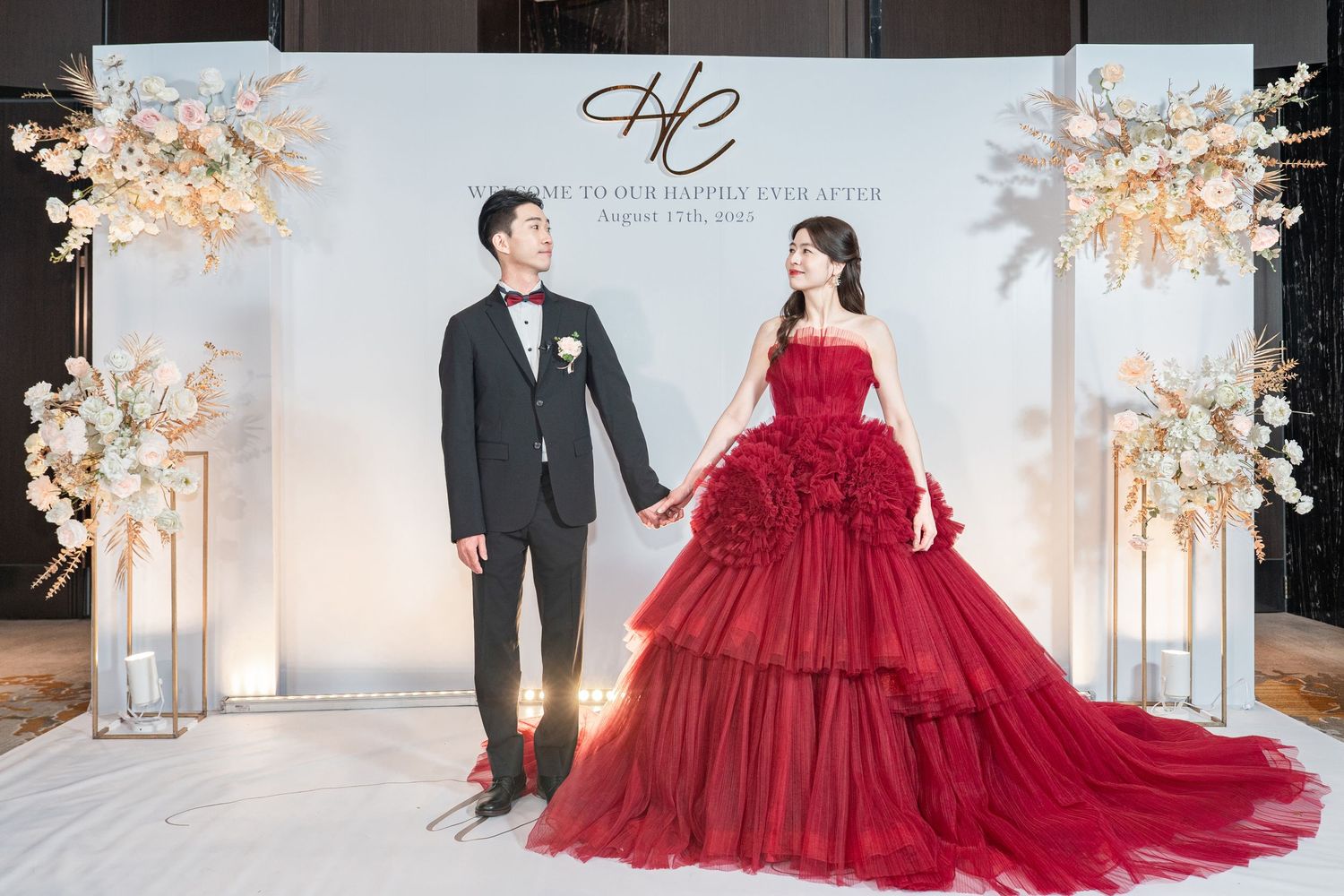 Elegant couple posing at wedding photoshoot with red ballgown and tuxedo against decorative white backdrop.