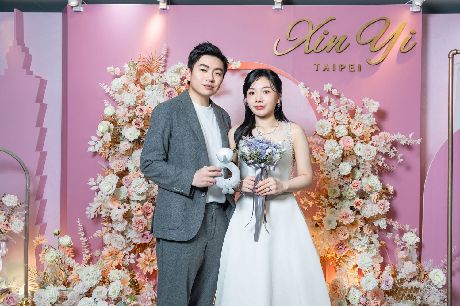 A couple in wedding attire poses in front of a pink floral backdrop at a formal event.