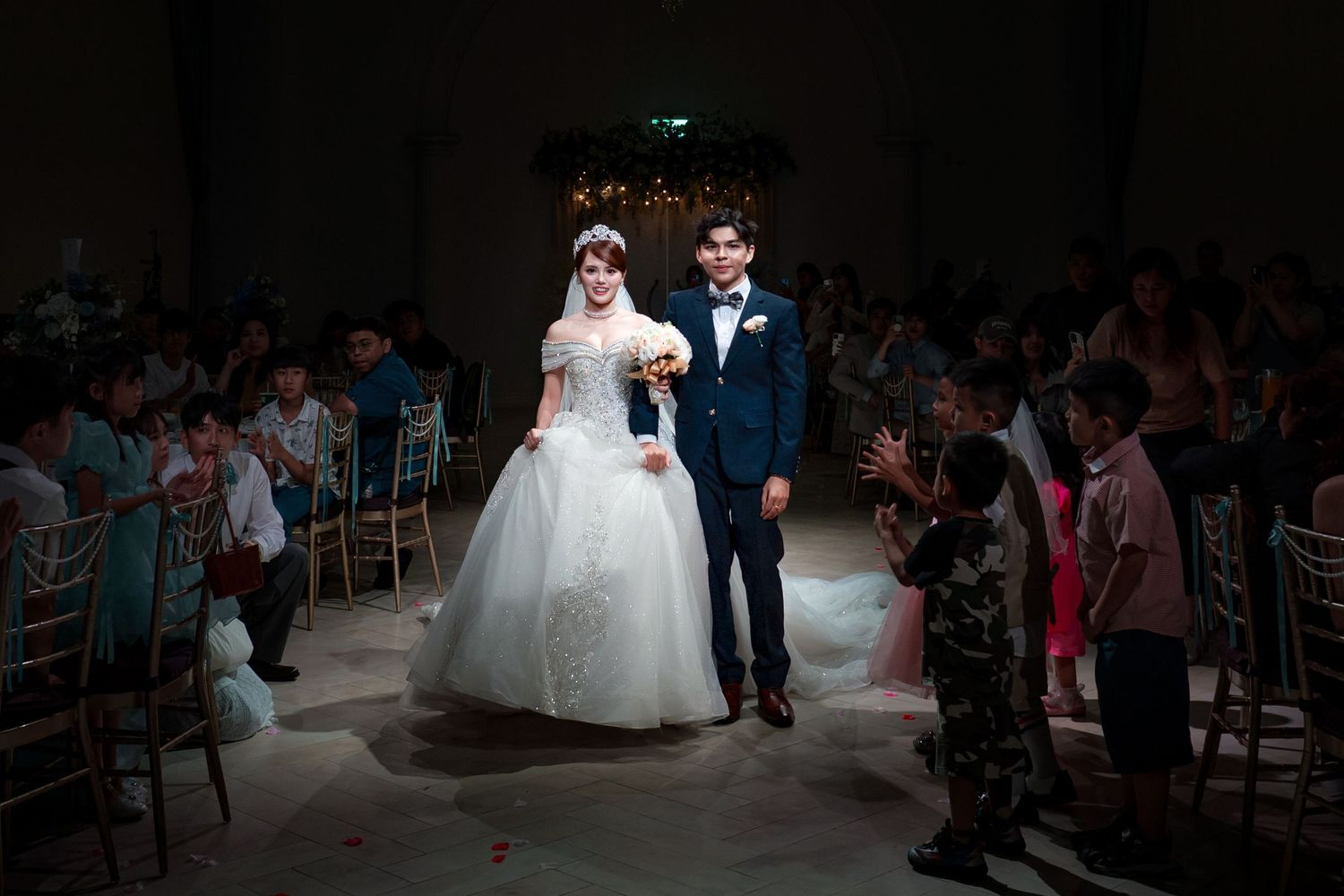 Bride in white ball gown and groom in navy suit walk down wedding aisle surrounded by seated guests.