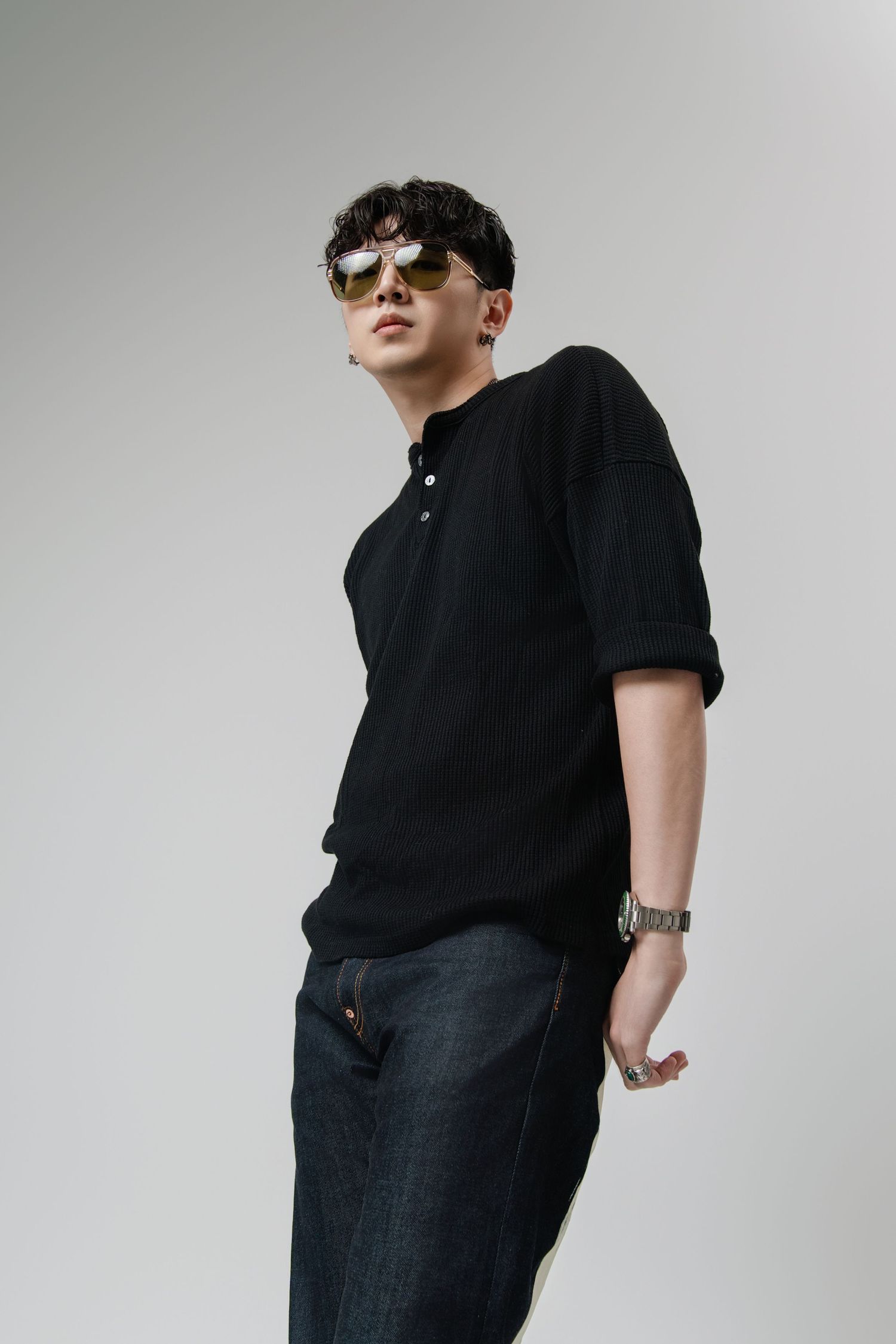 Young man in black henley shirt, dark jeans, sunglasses, and watch posing against gray background.