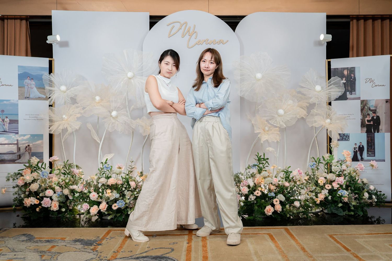 Two women pose elegantly in front of a floral wedding backdrop with white blooms and soft pastel decor.