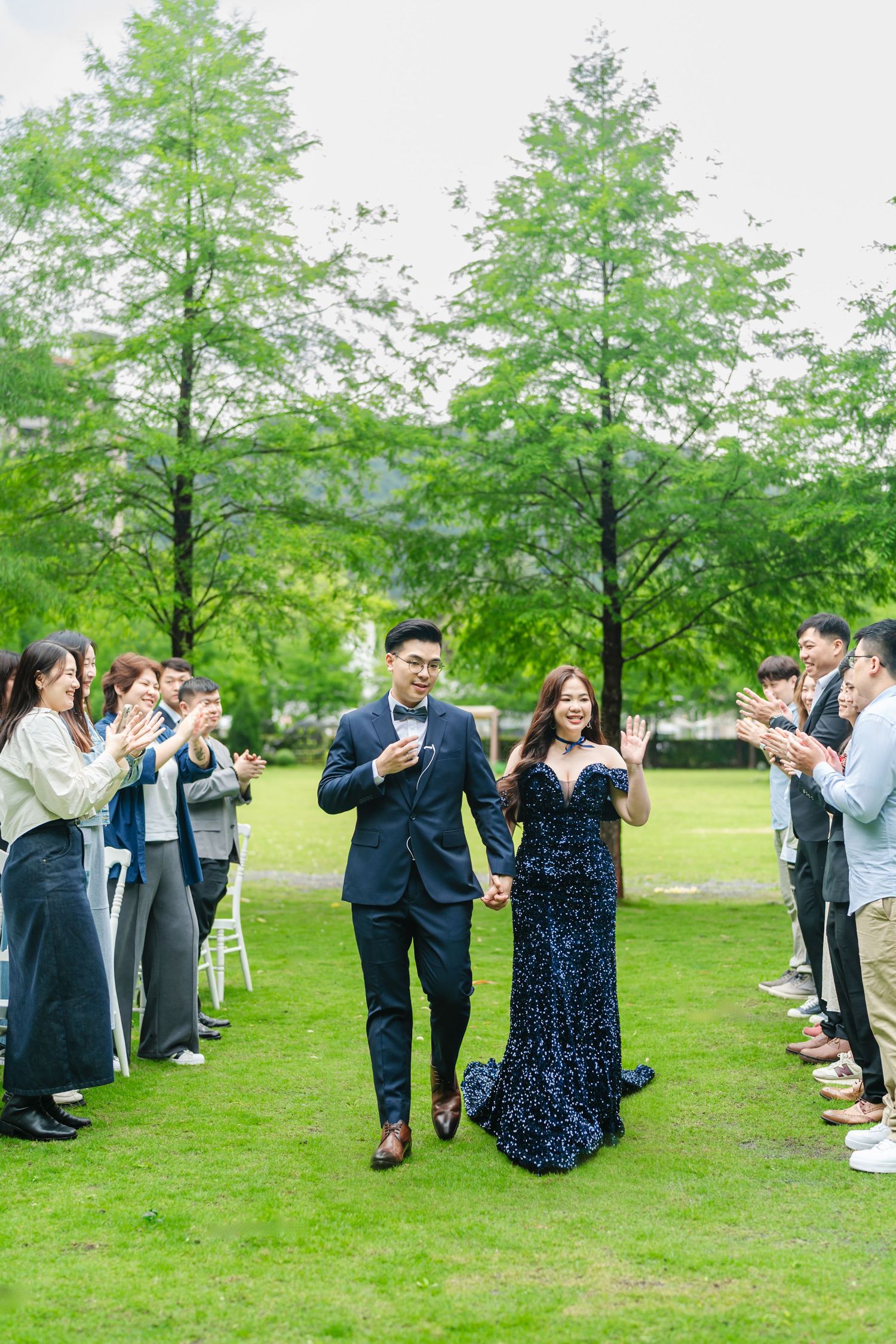A couple and guests celebrate on a green lawn lined with tall trees at an outdoor event.