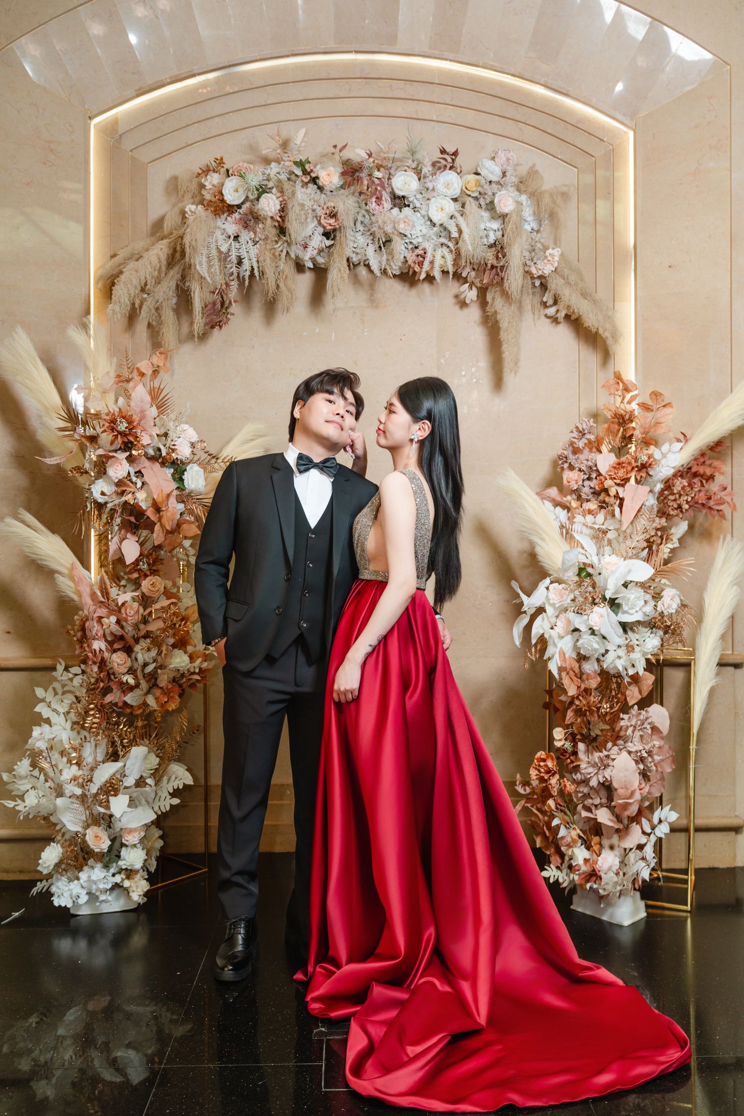 Elegant couple poses under floral archway with ornate decor, bride wearing stunning red gown and groom in classic tuxedo.