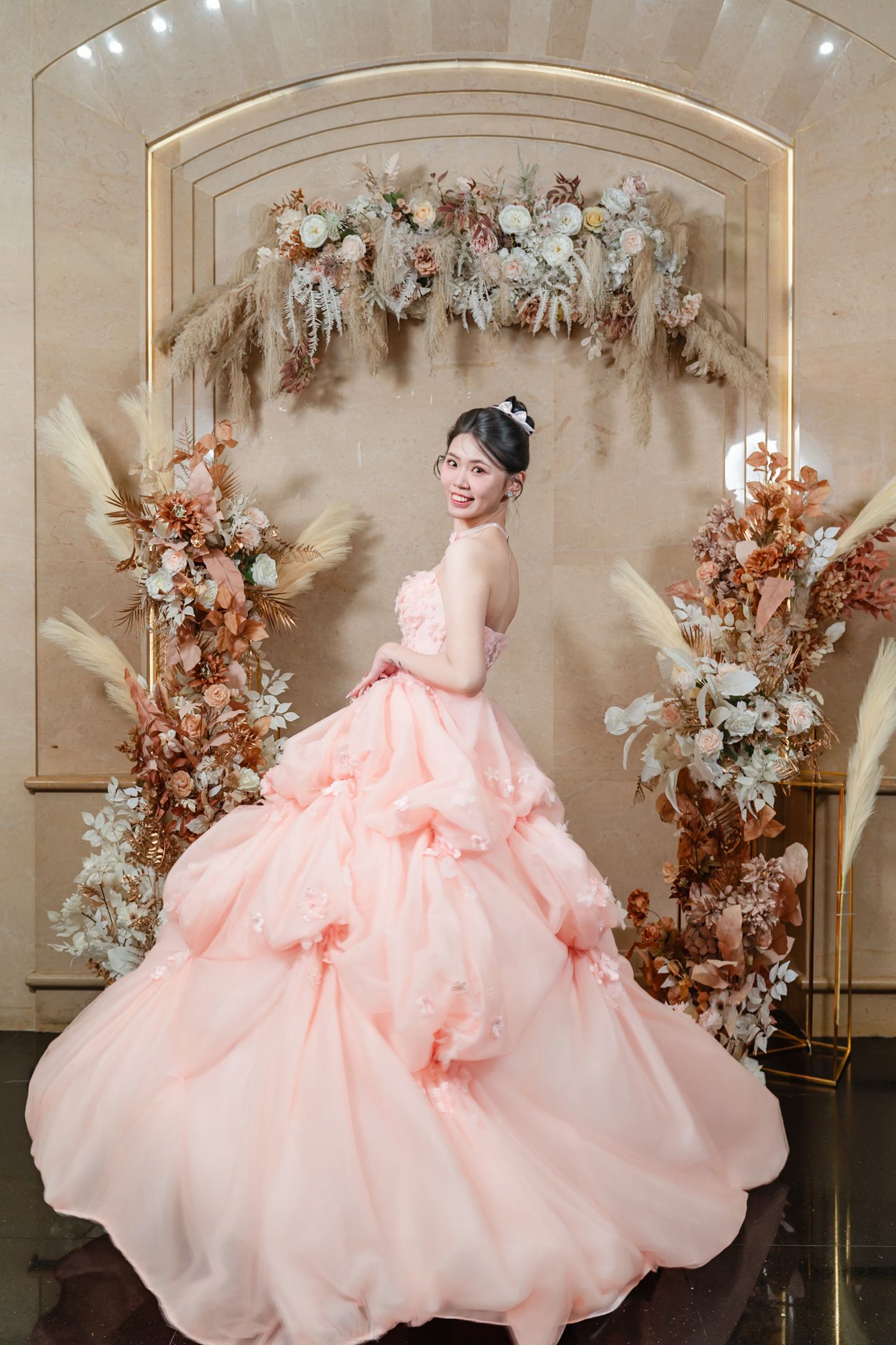 An elegant peach ballgown with floral decor against an arch adorned with dried flowers and white blooms in a studio setting.