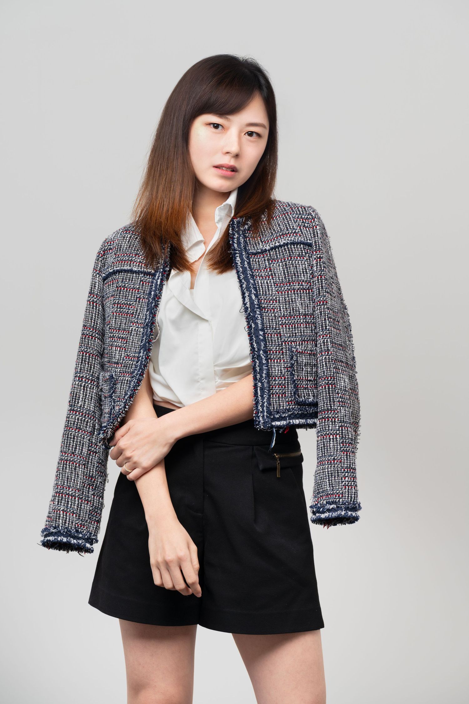 A woman poses in a tweed jacket, white blouse and black mini skirt against a gray background.